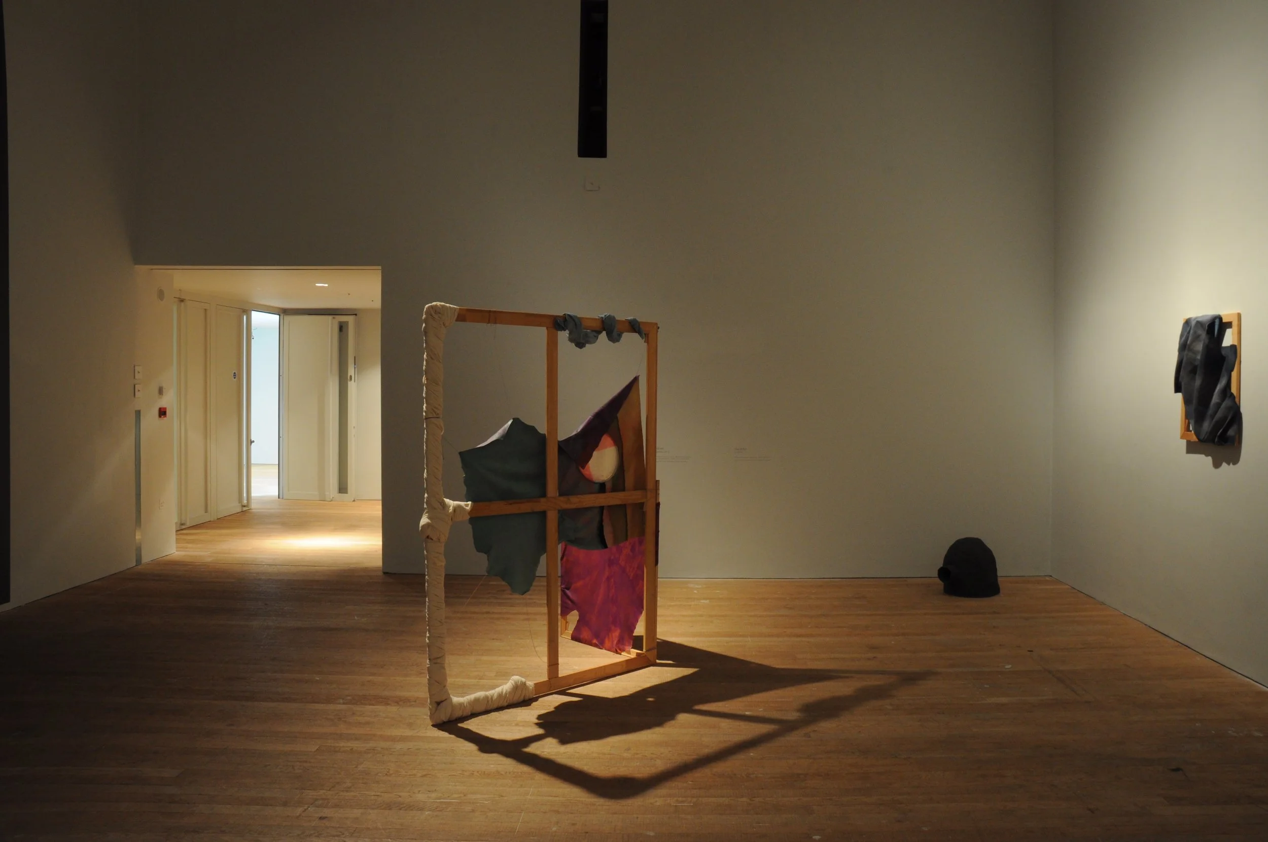 An art gallery with a wooden sculpture in the center, consisting of fabric and wood, with a shadow cast on the wooden floor. There are other art pieces on the walls and a black object on the floor.