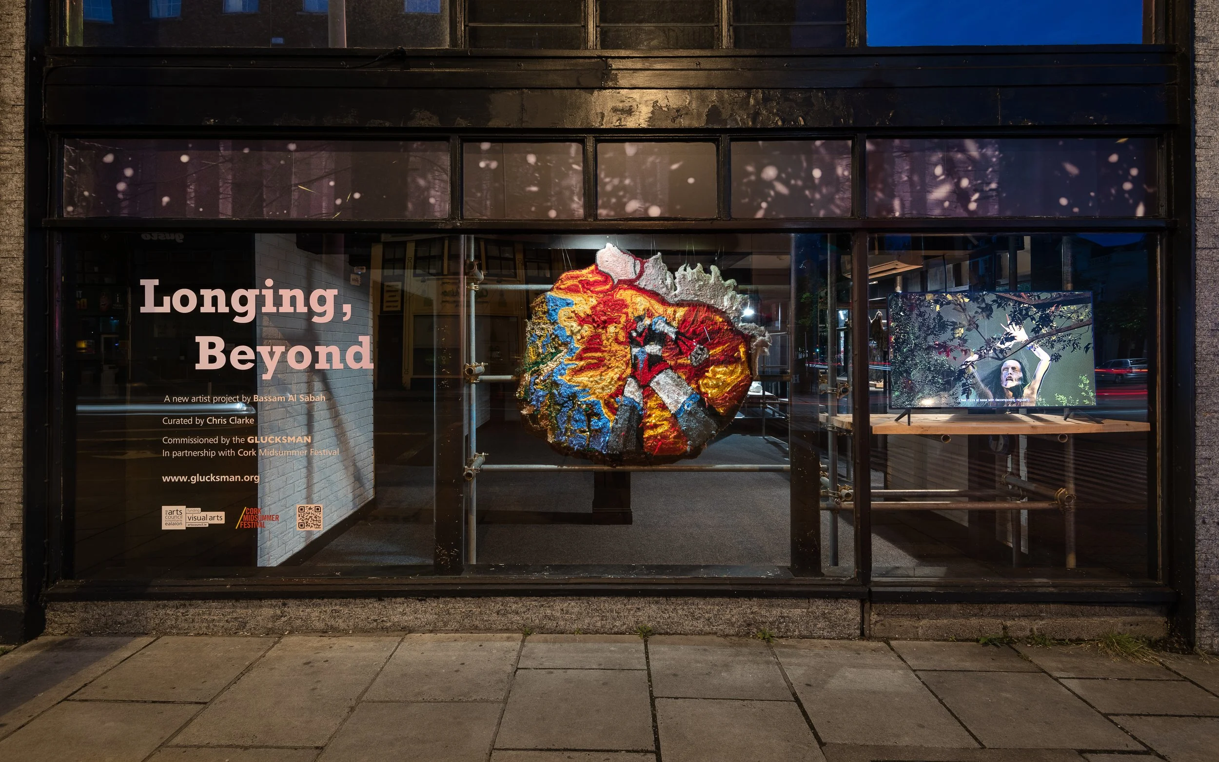 Gallery window display with poster titled 'Longing, Beyond' showing a colorful round textile art piece and a small monitor displaying a video of a person dancing outdoors.