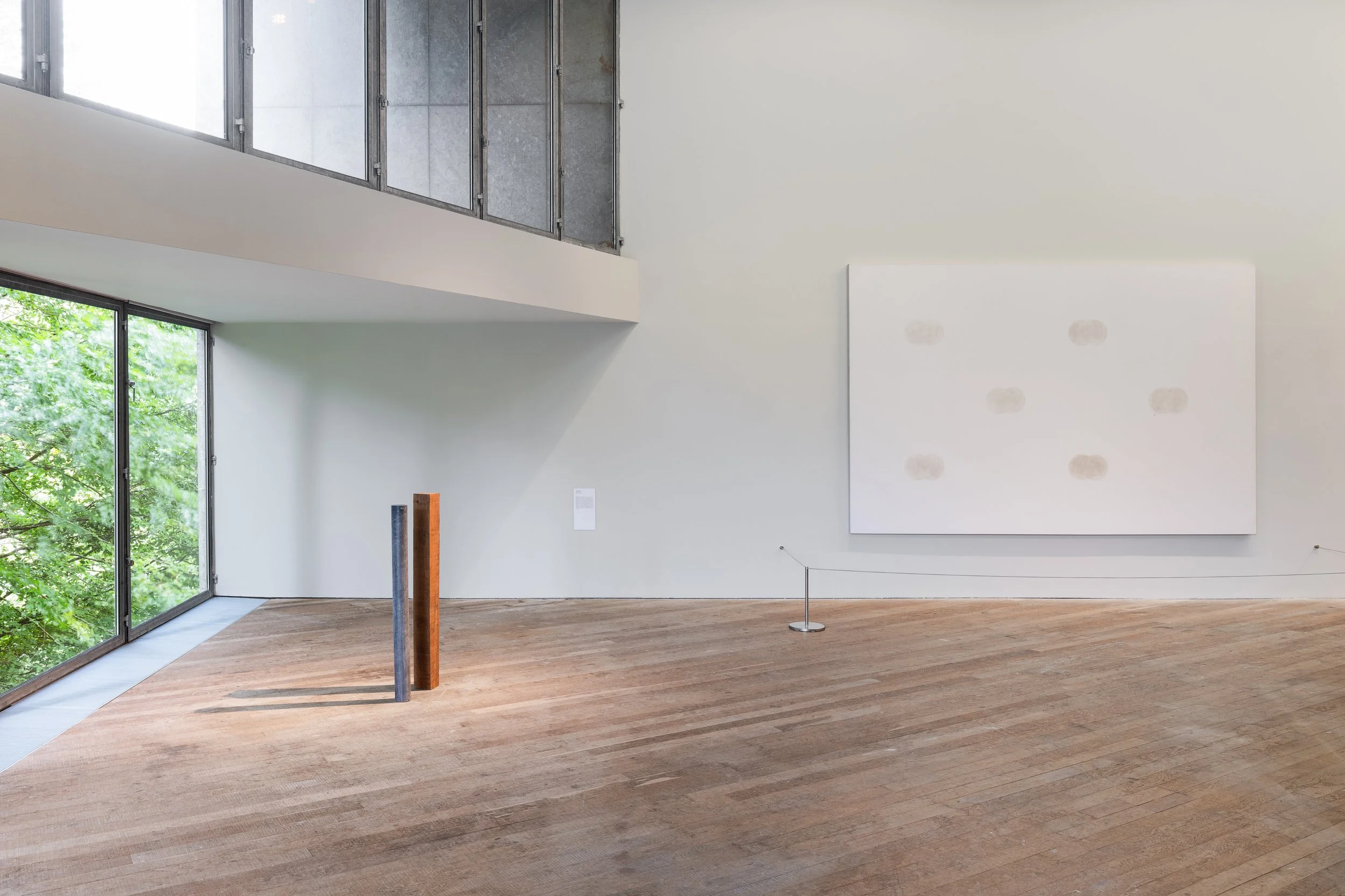 Contemporary art gallery with wooden flooring, white walls, large window revealing green trees outside, abstract artwork on wall, and three vertical sculptures.