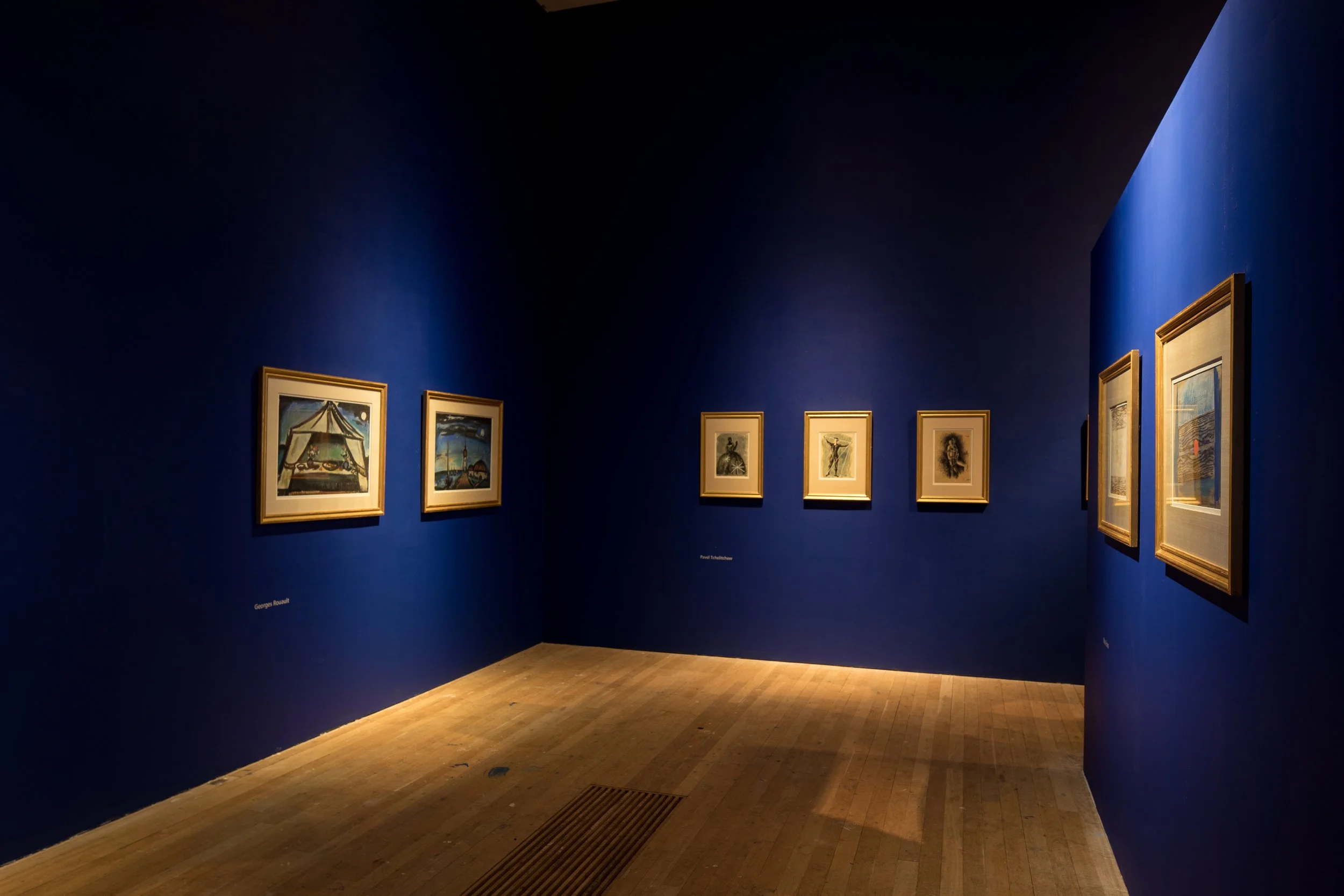 An art gallery with blue walls displaying framed paintings in a well-lit room with a wooden floor.