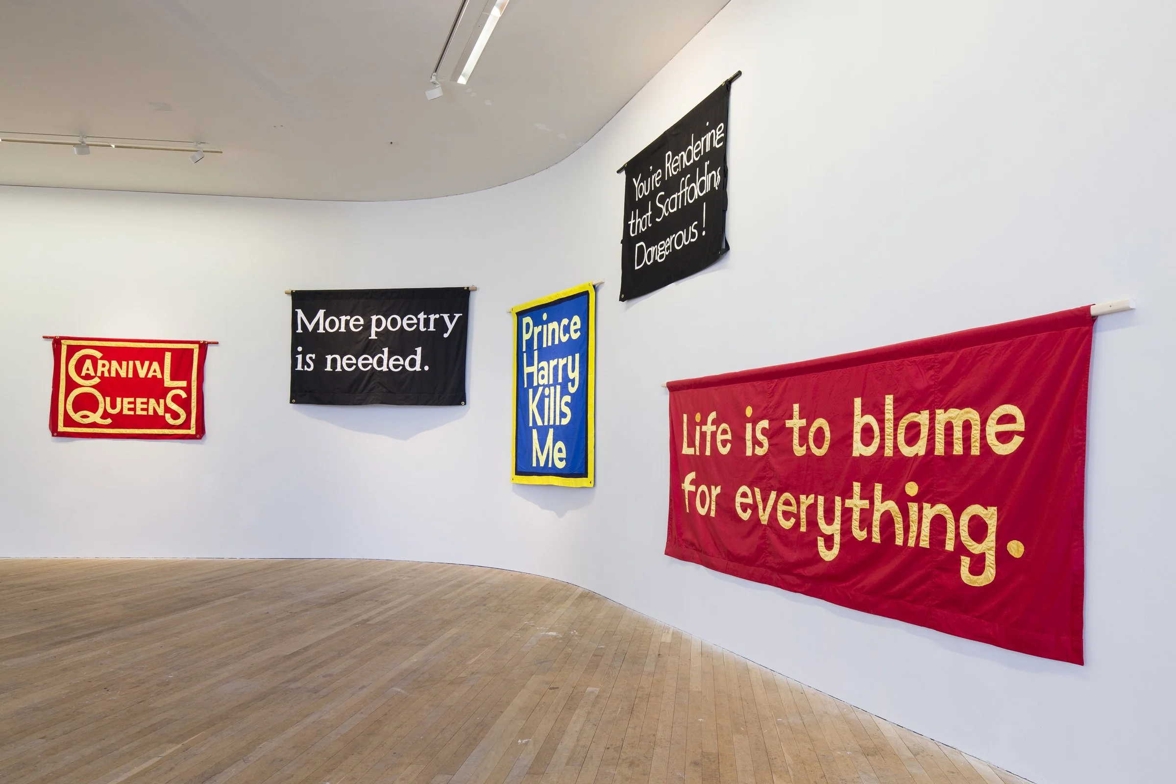 Gallery wall displaying six fabric banners with various text messages in an art gallery space with wooden floor and white walls.