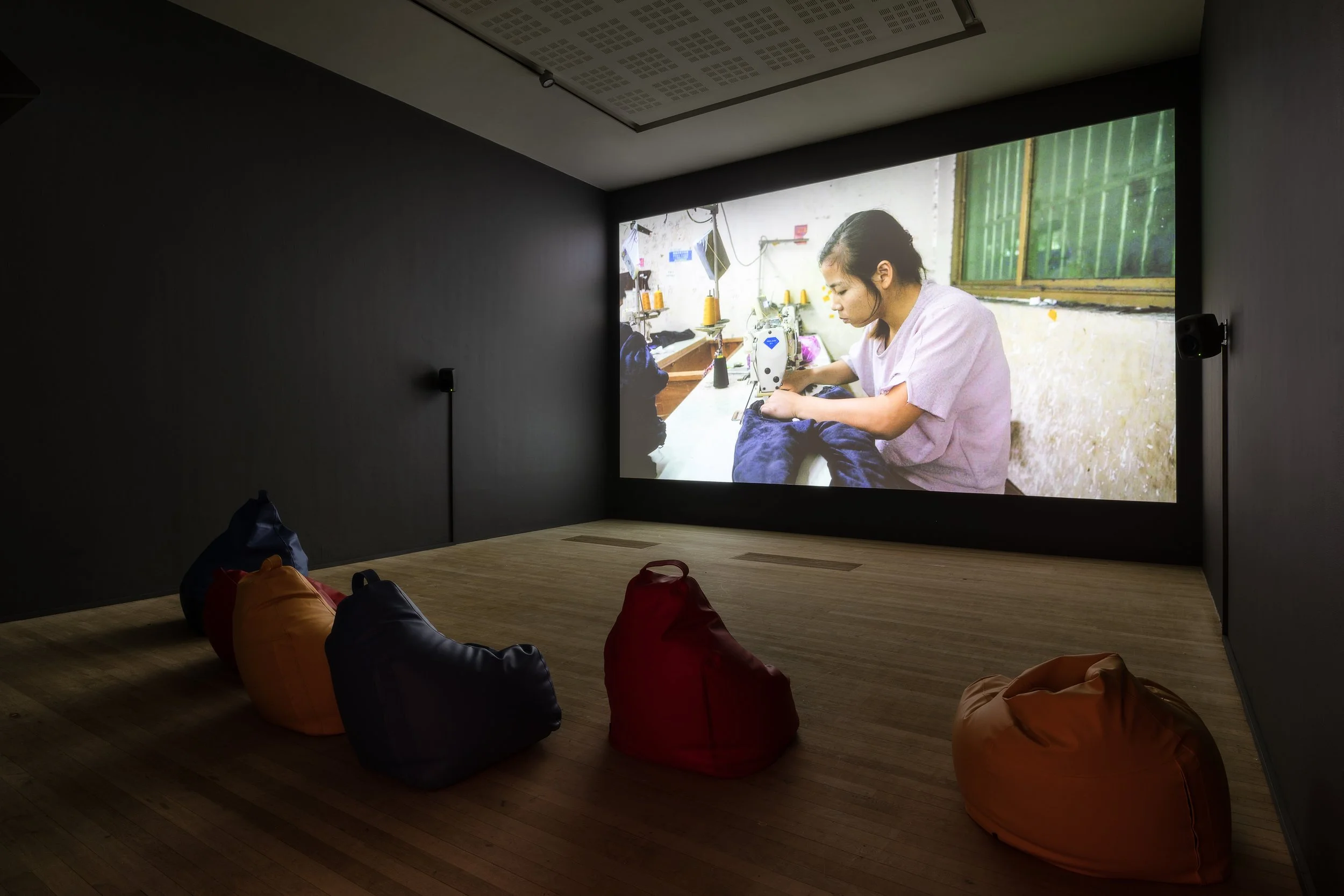 A gallery with bean bag chairs on the floor facing a large screen displaying a woman sewing fabric in a workshop.