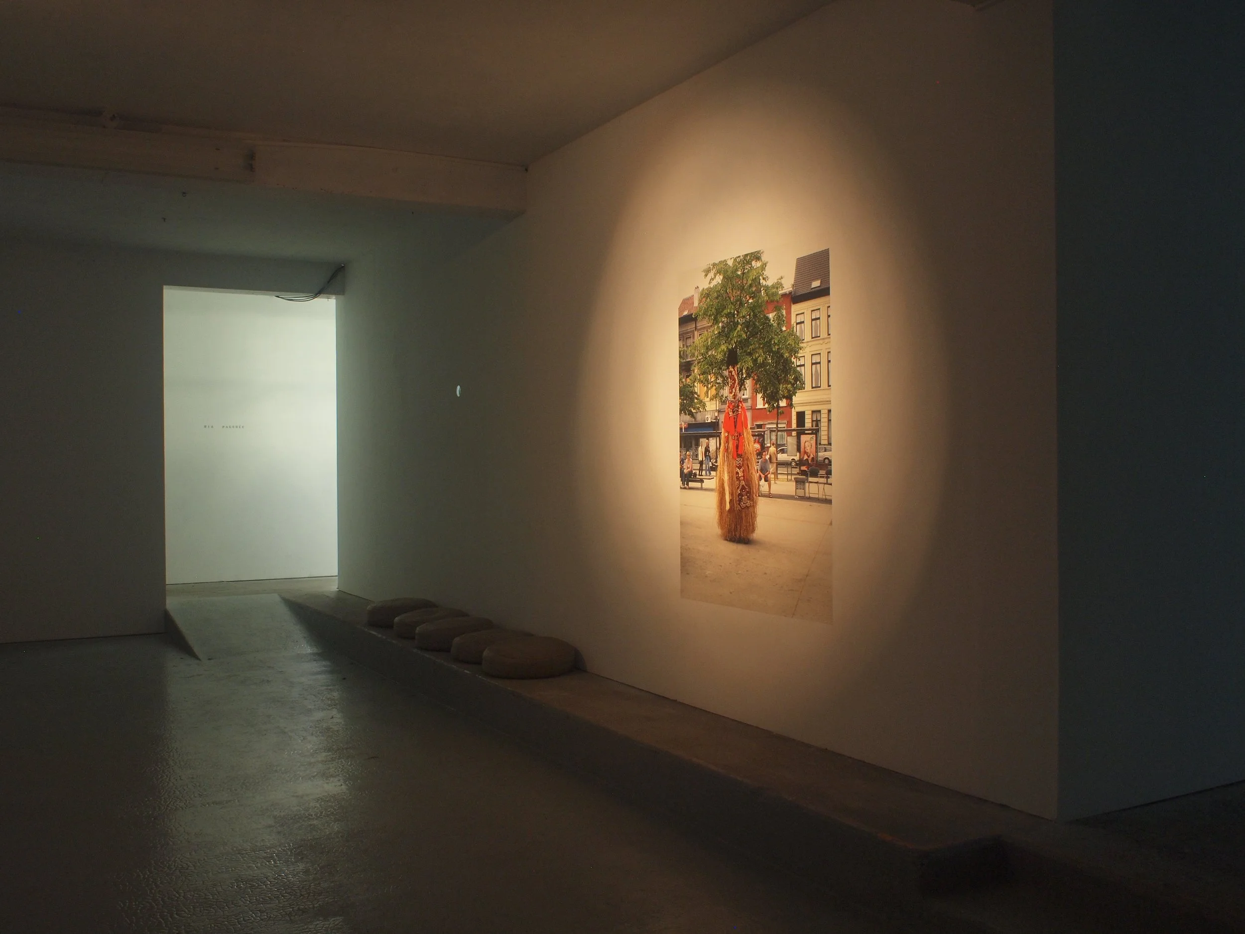 An art gallery with a photograph of a tree and an orange statue in an urban setting, illuminated by a spotlight, with a row of round cushions on the floor nearby.