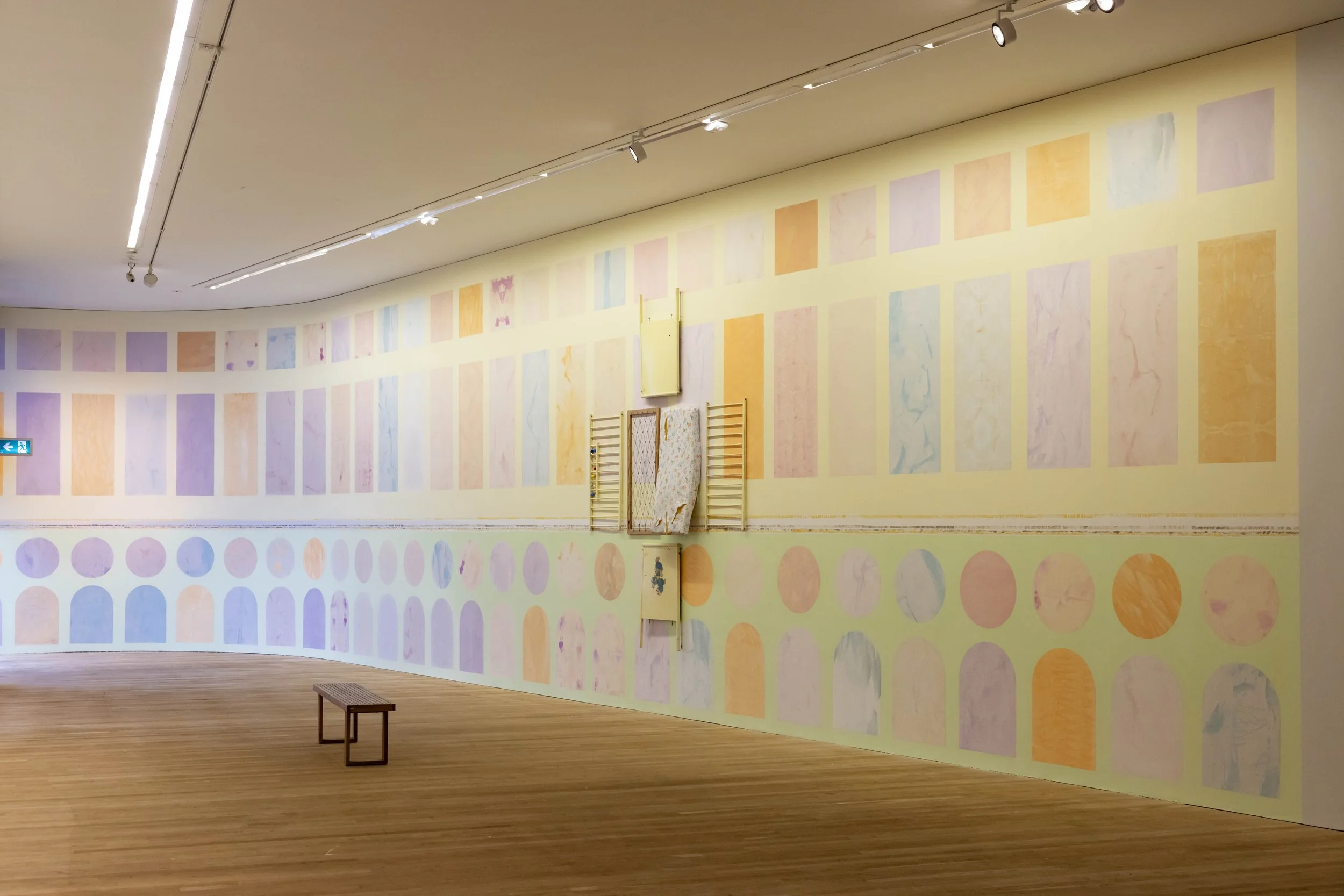 An art gallery with a large pastel-colored wall display of various rectangular and circular watercolor paintings, a wooden bench, and ceiling lights.