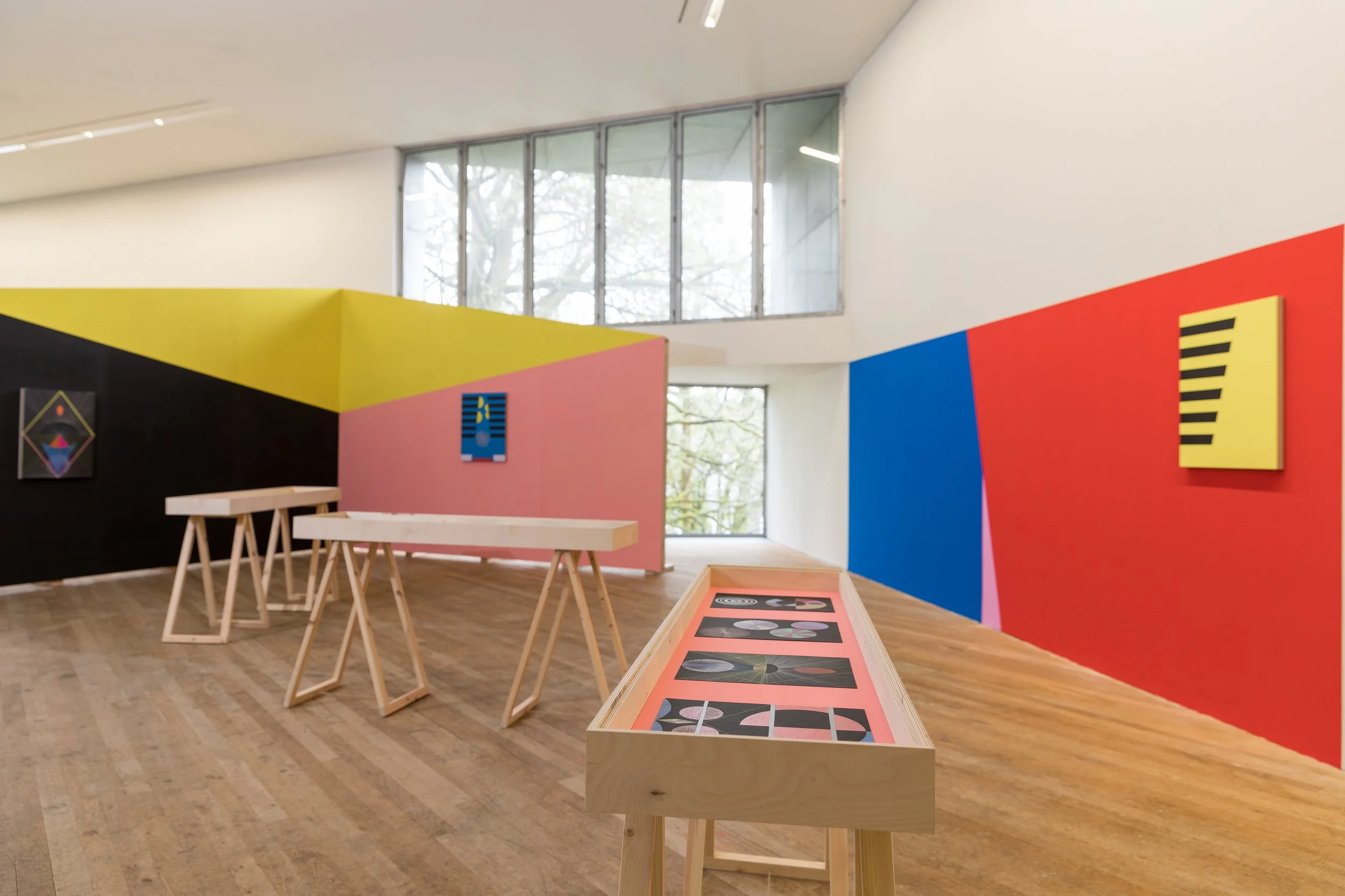 An art gallery with colorful walls, displaying geometric paintings, and wooden display tables on a hardwood floor.