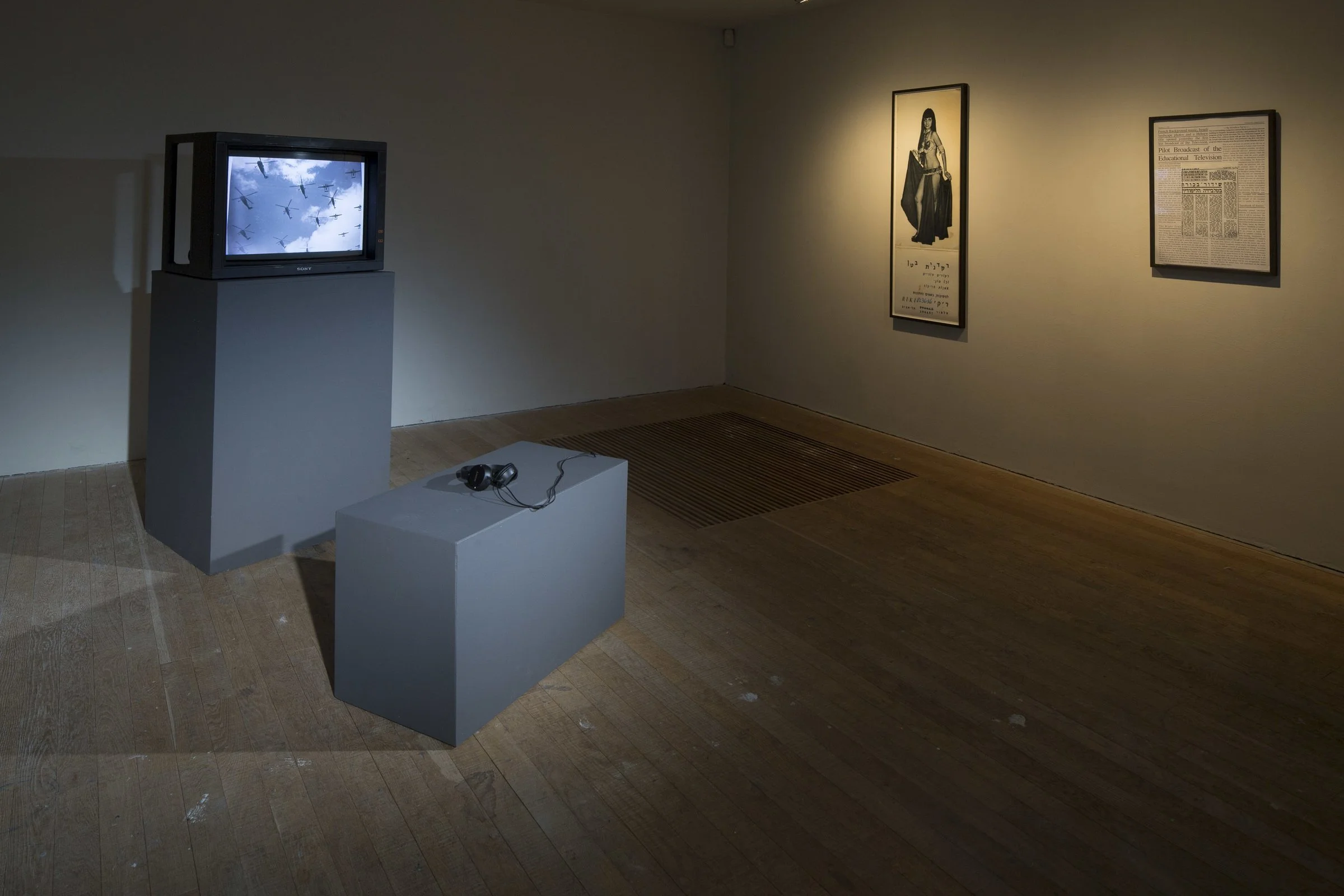 An art gallery display with a vintage television showing a video with flying toy airplanes, a pair of headphones on a pedestal, and framed artwork and a newspaper on the wall.