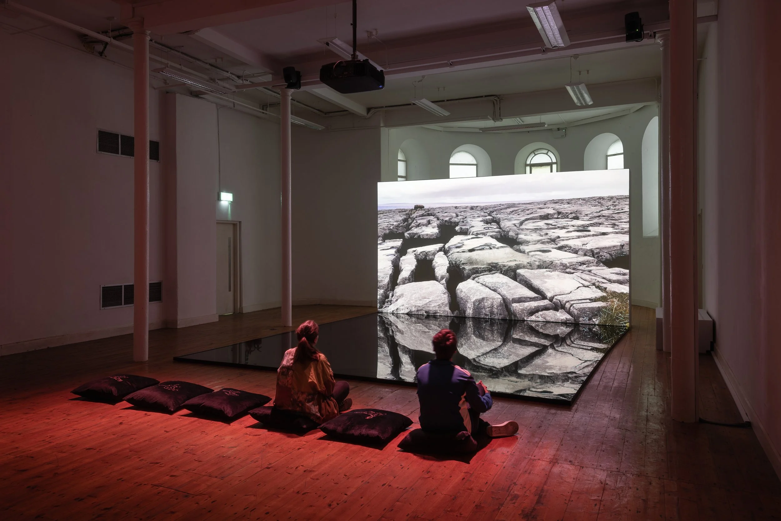 Two people sitting on floor cushions watching a large digital display of a rocky landscape in an art gallery.