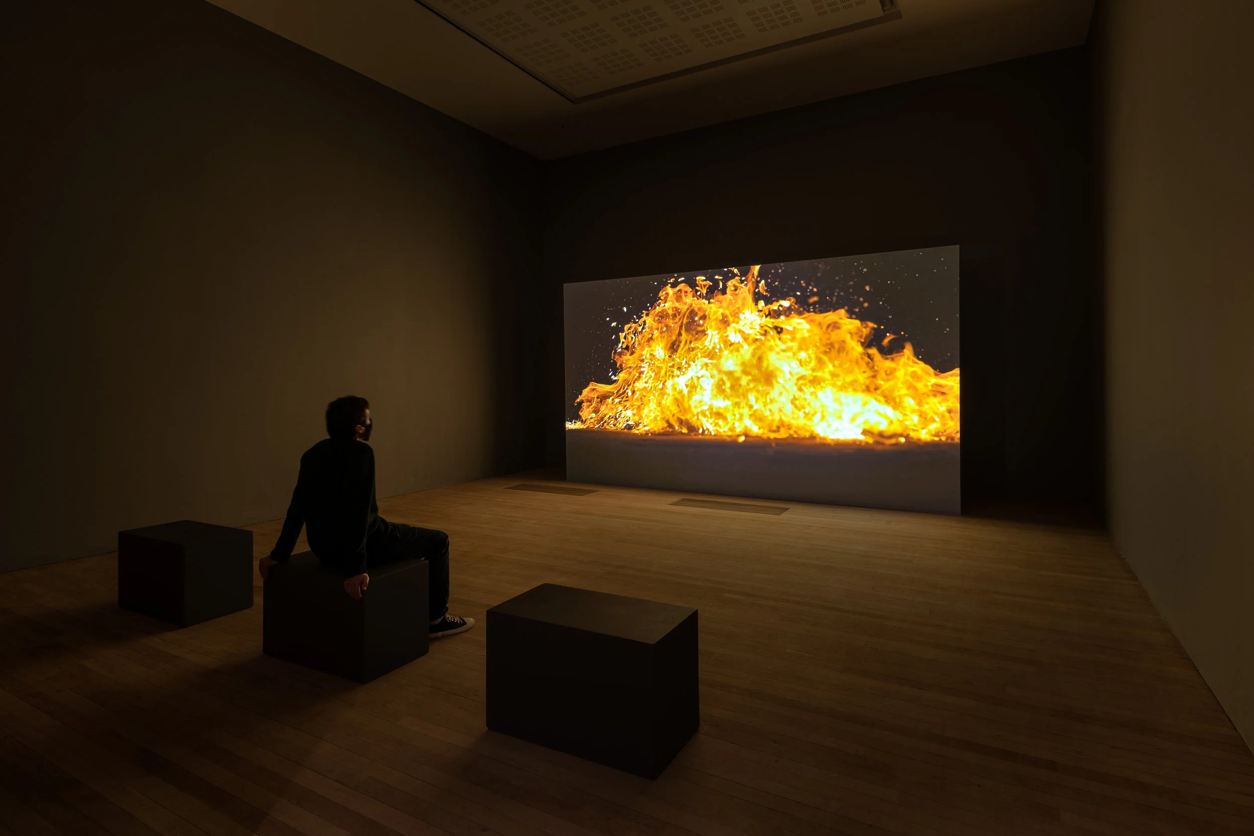 Person sitting on a cube watching a large screen displaying an explosion or fire in a dimly lit room.