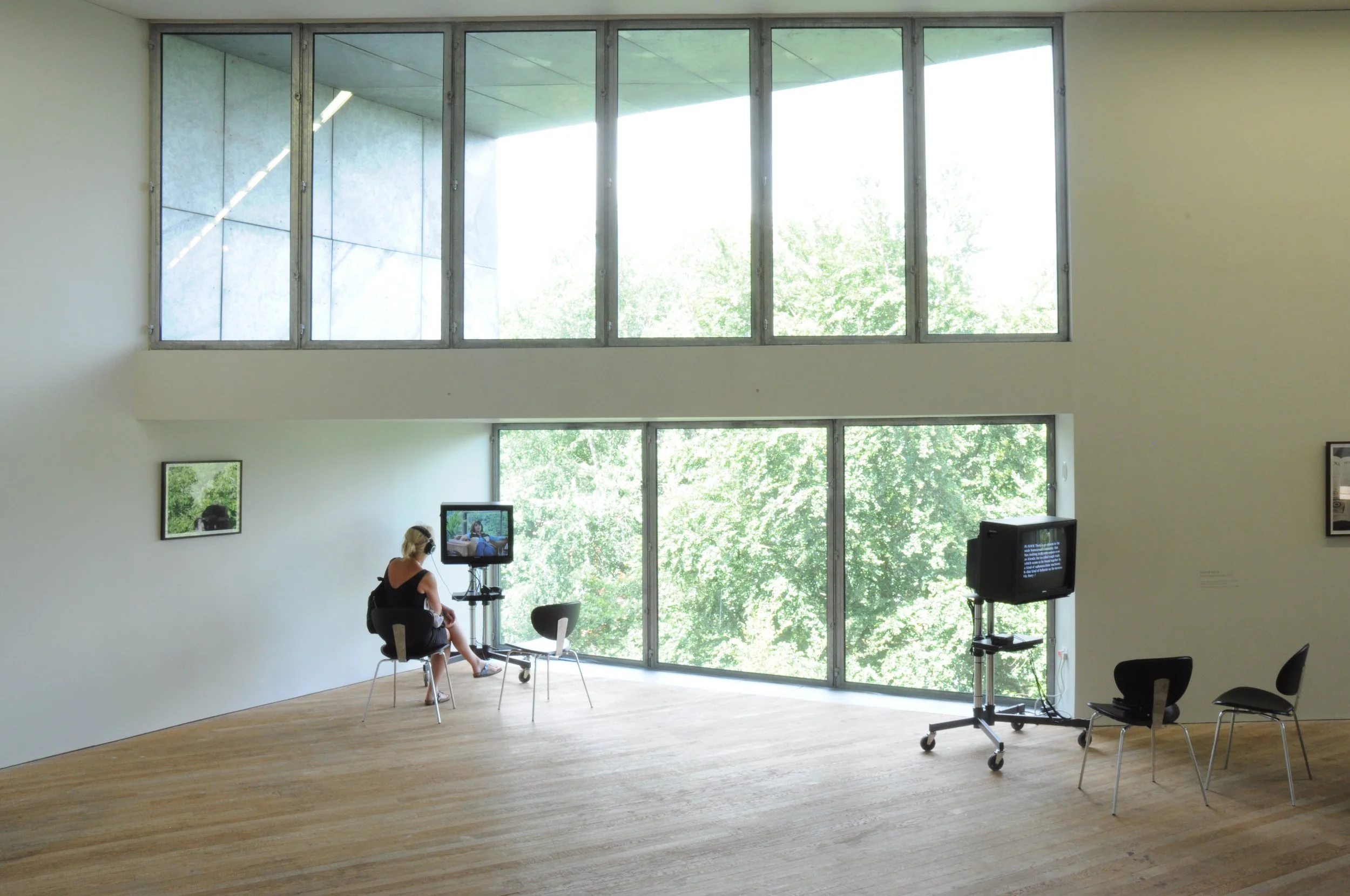 A woman sitting on a chair in an art gallery, looking at a monitor on a stand. The picture is minimalist, with large windows showing greenery outside, wooden flooring, and a few chairs, one with a TV on a stand.