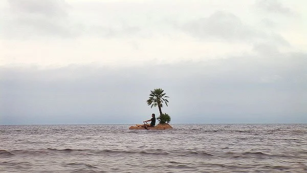 A small island with two palm trees and a person sitting on the rocks, surrounded by water under a cloudy sky.