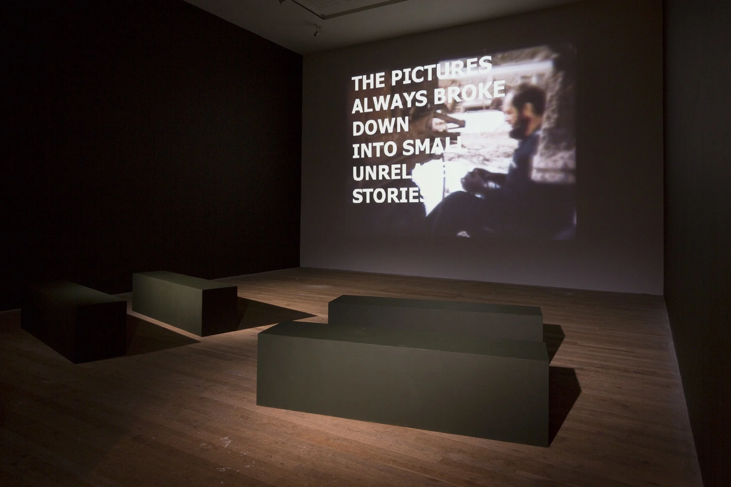 An art installation or exhibit featuring a large screen displaying a woman sitting outdoors and a quote that reads, 'The pictures always broke down into small unreliable stories.' The room has a dark background with a wooden floor.