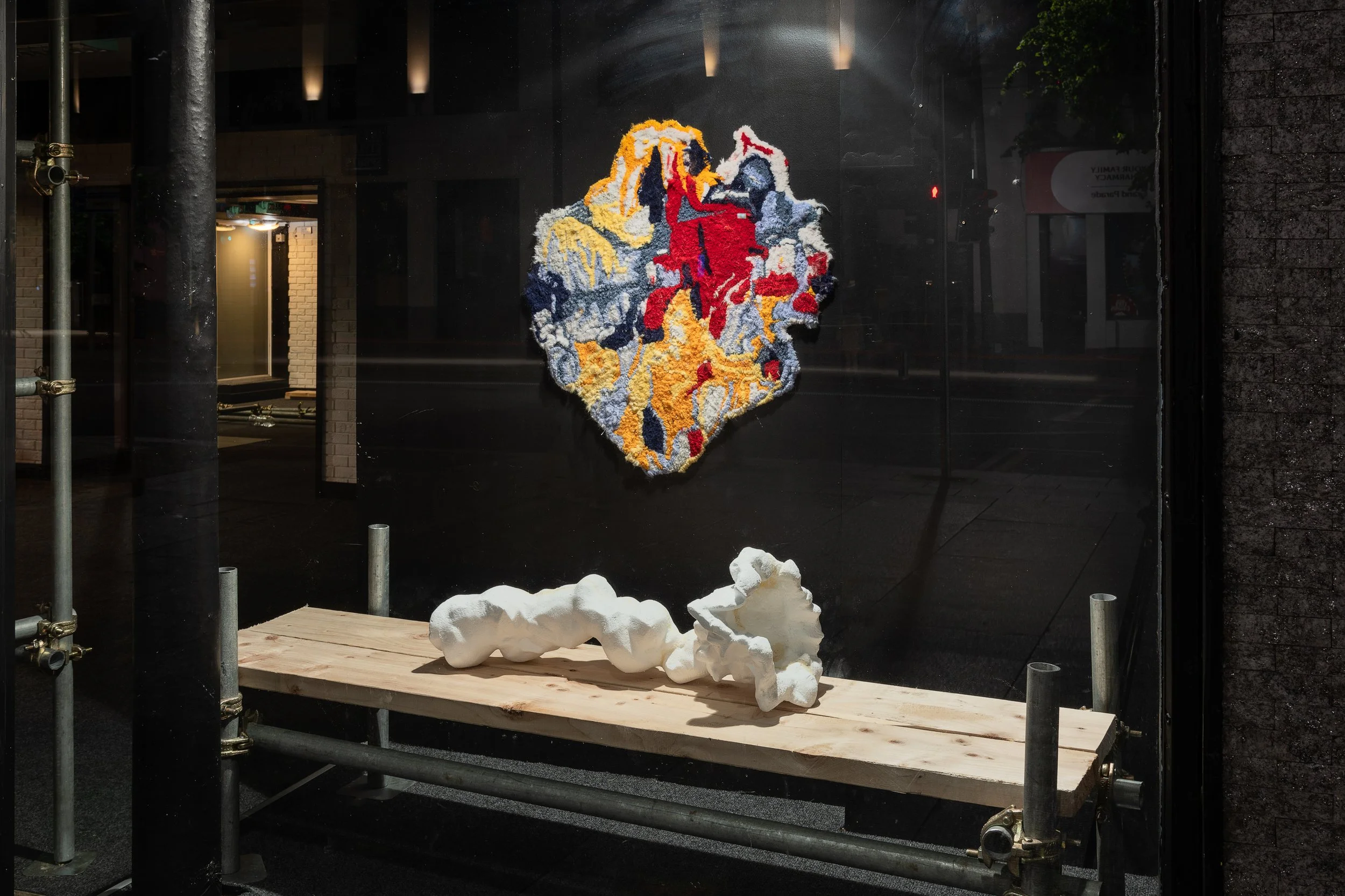 An art display on a storefront window featuring a colorful, textured lion's head made from yarn or fabric, mounted on a black background. Below it, on a wooden platform, are two abstract white sculptures resembling cloud formations.