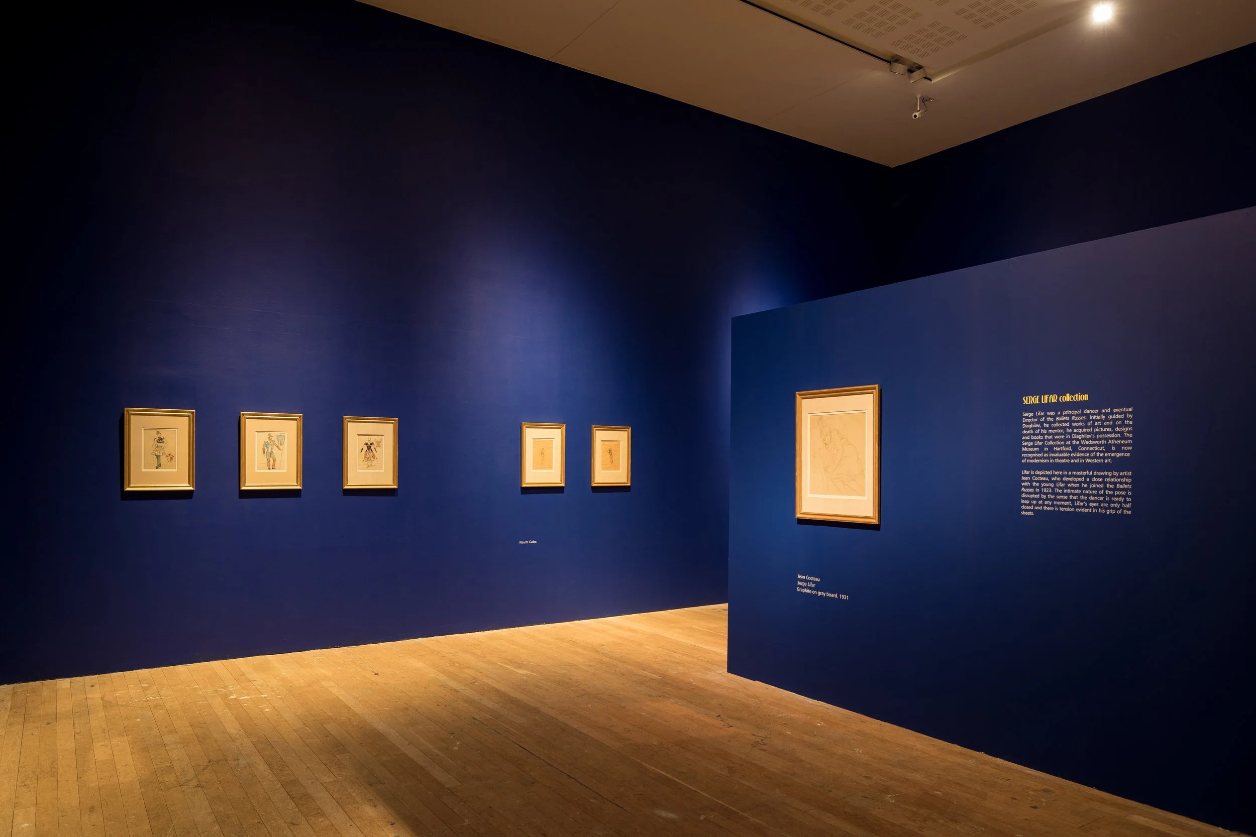 An art gallery with blue walls displaying five framed sketches of ballet dancers and a large framed graph on the right wall. There is a shadow on the wall and a wooden floor.
