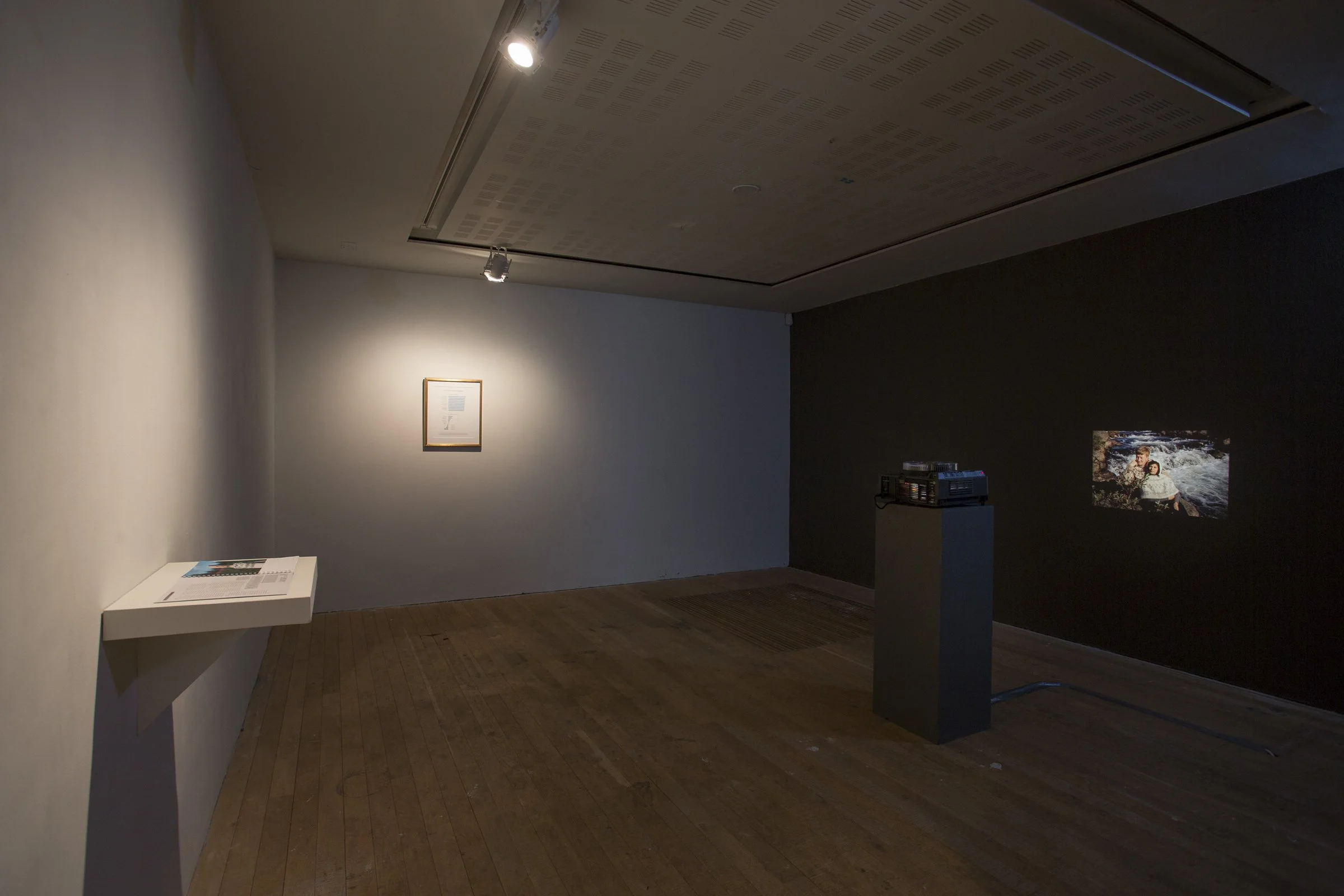 An art gallery room with a framed picture on the wall illuminated by a spotlight, a pedestal with a stack of media items, a small display on a ledge, and a projected photograph on the dark wall.