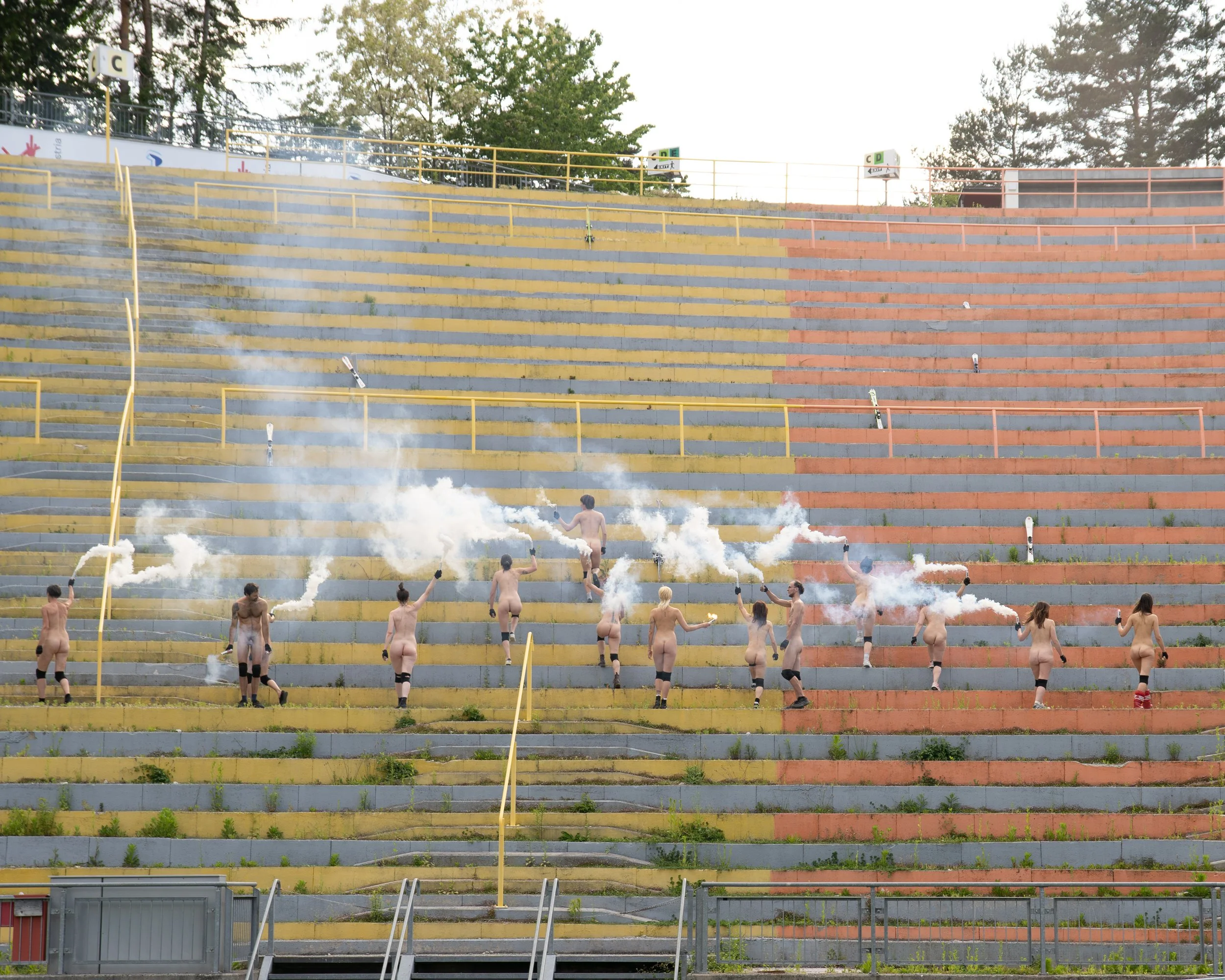 People marching up stadium bleachers while holding smoke bombs in an empty outdoor stadium.