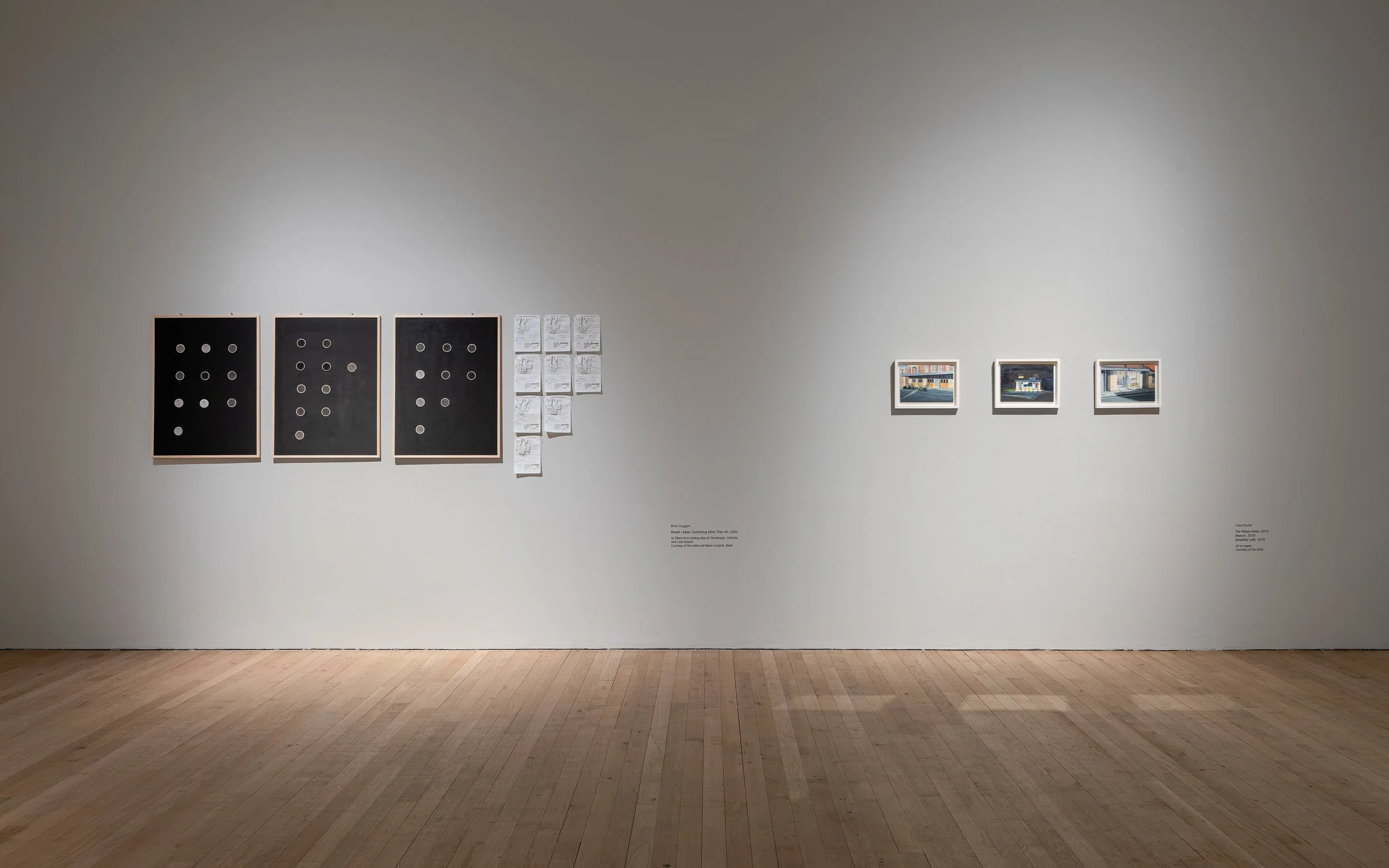 An art gallery wall displaying three framed black-and-white abstract artworks with circular shapes on the left, and three smaller framed colorful photographs on the right. There are also several sheets of paper with notes or sketches pinned to the wa