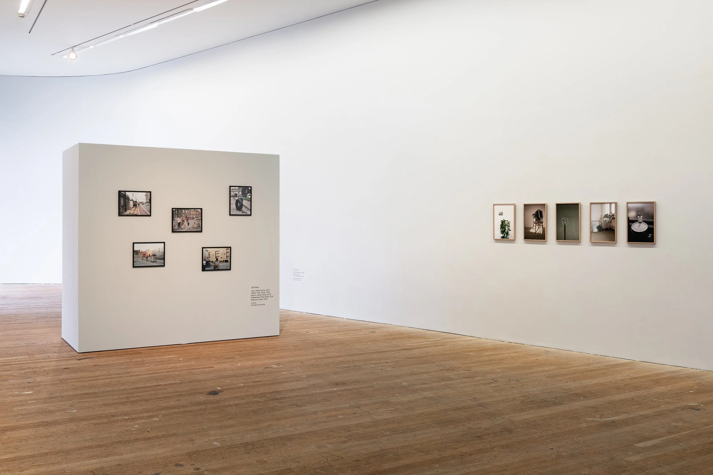 Empty art gallery with a white wall featuring six small framed photographs and a freestanding white wall with six more photographs. Wooden floor and minimal lighting.
