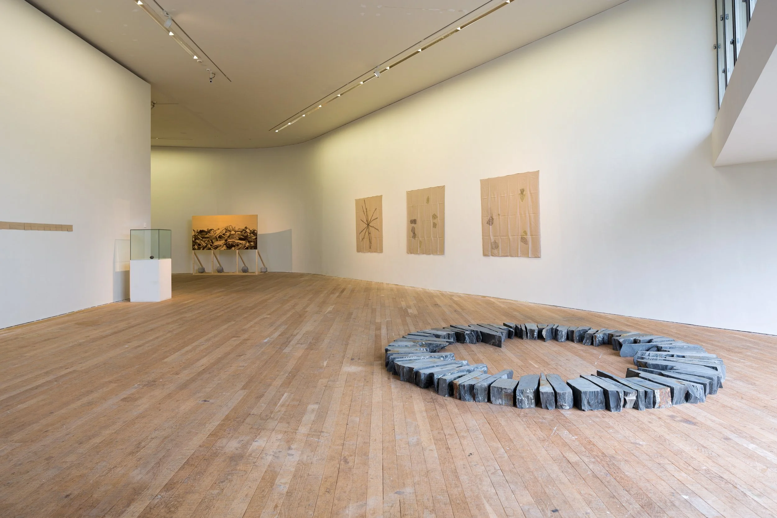 Art gallery with wooden flooring, beige walls, and natural light. Features a large stone art piece in a circle on the floor, paper artworks on the wall, and display cases.
