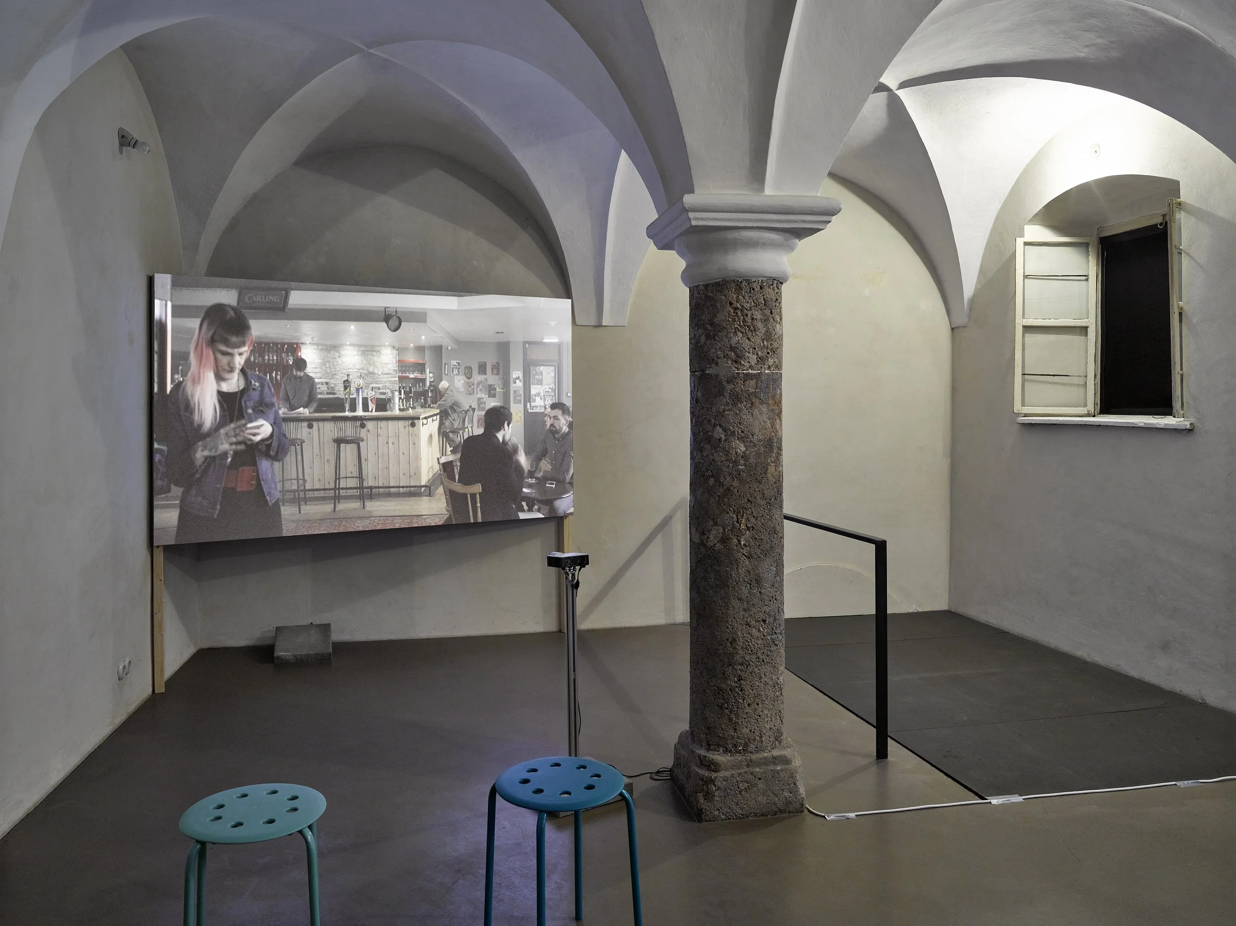 An art installation or exhibition space with a stone column, arched ceiling, small window, and chairs in foreground. A large digital screen displays a black-and-white photo of a bar scene with people socializing.