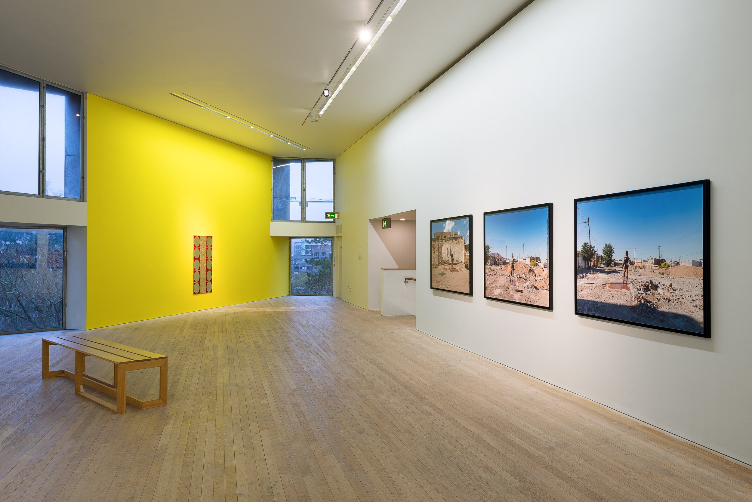 Contemporary art gallery with three framed photographs on white wall, yellow accent wall, wooden bench, large windows, and track lighting.