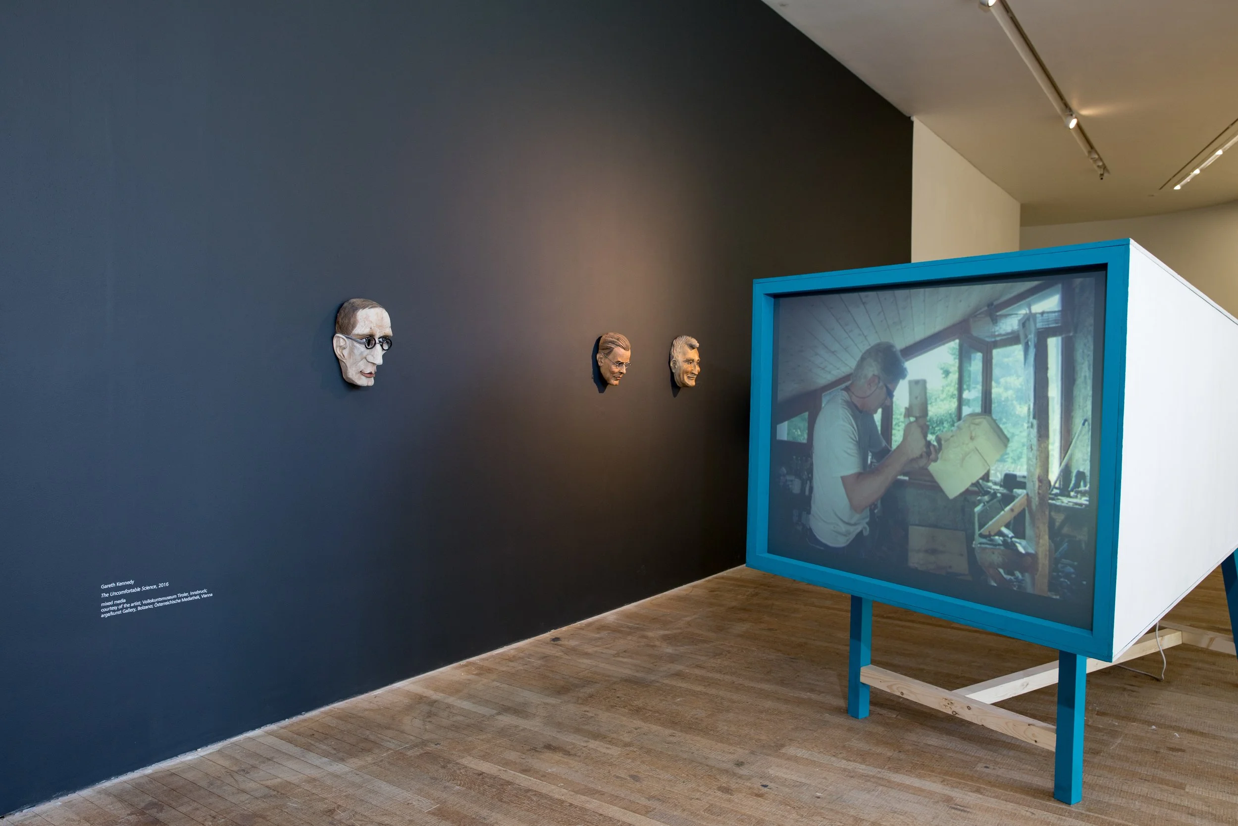 An art exhibit featuring four three-dimensional sculpted heads of men mounted on a dark-colored wall, and a large display screen framed in blue showing a man sketching at an easel in a bright room.