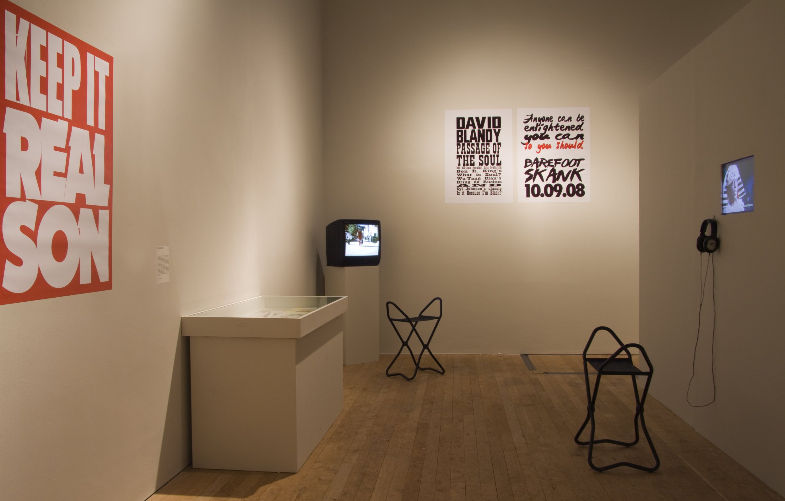 An art exhibit room with posters on white walls, a small CRT TV on a pedestal showing a scene, two black wire chairs, a wall-mounted screen with headphones, and a large red and white poster reading "Keep It Real Son."