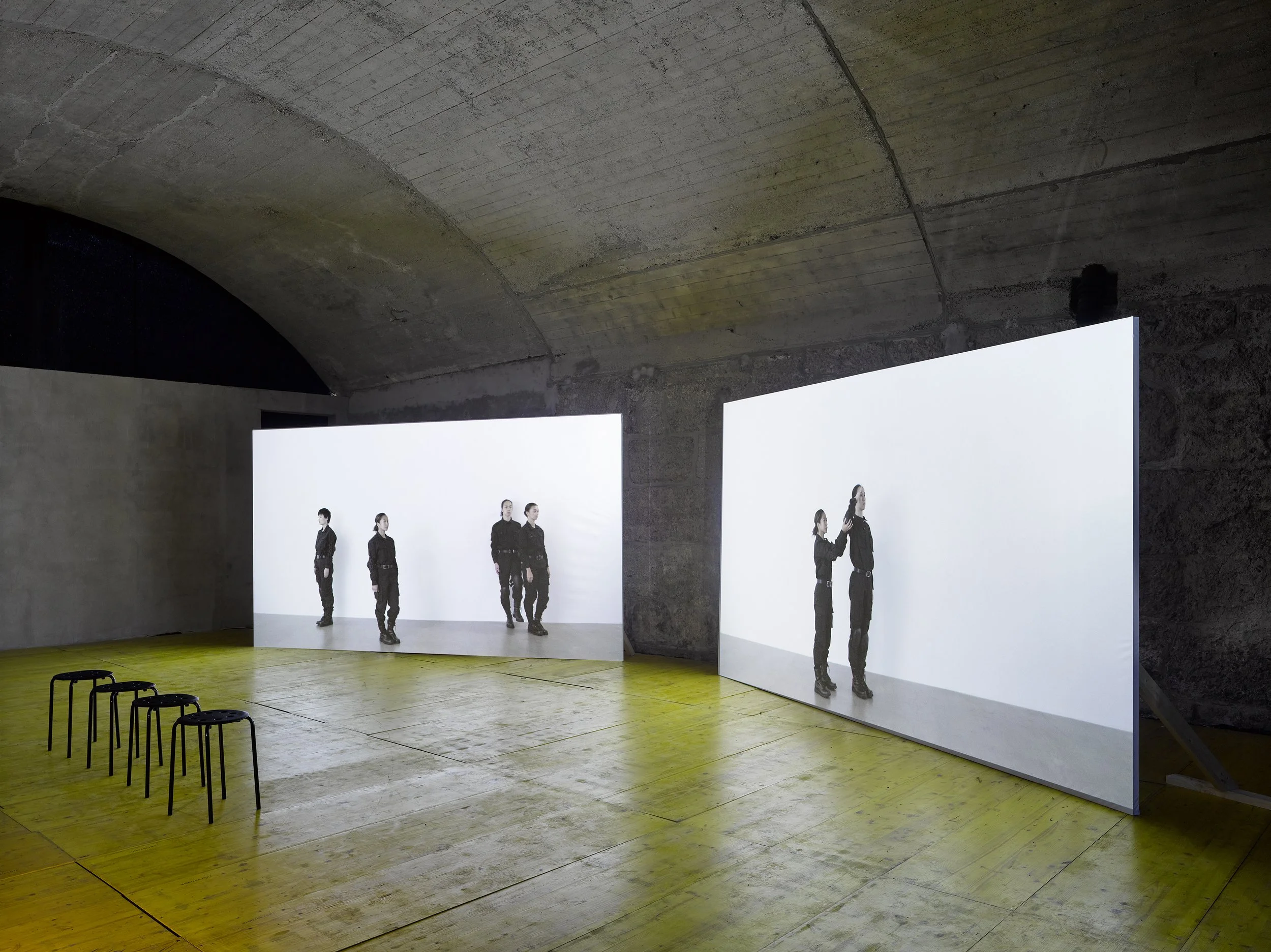 An art installation with two large screens displaying black-and-white photos of people in uniform, set inside an underground concrete space with a wooden floor and four black chairs.