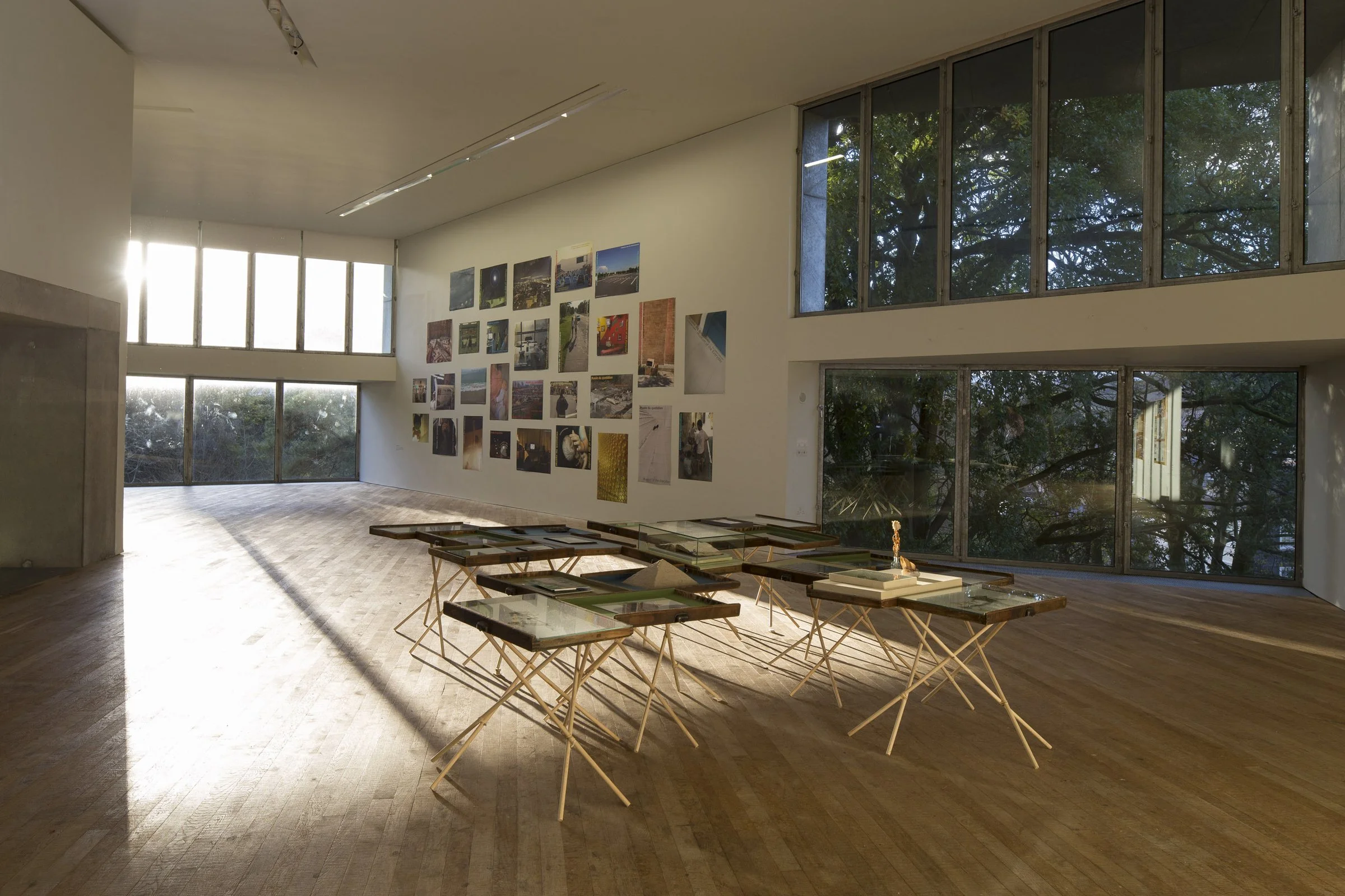 An art gallery with wooden floors and large wall windows showing trees outside. The gallery has a wall with multiple photographs and art displays. There are several glass-topped tables arranged on the floor, with some displaying books, papers, and a 