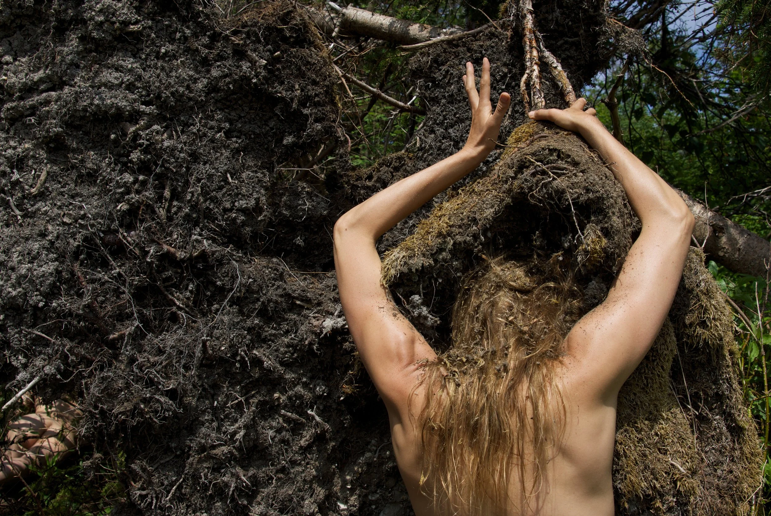 A woman with long hair leaning against a large tree with exposed roots, surrounded by green foliage.