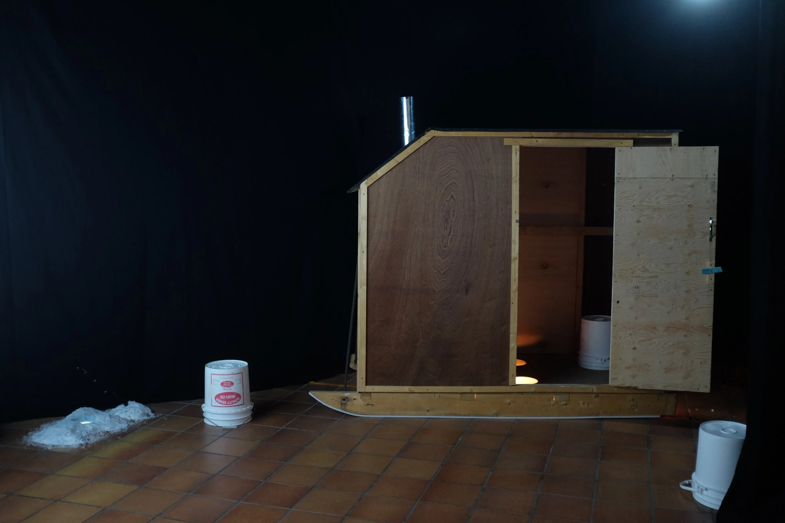 Unfinished wooden structure on a tiled floor with buckets and insulation foam nearby, against a black background.