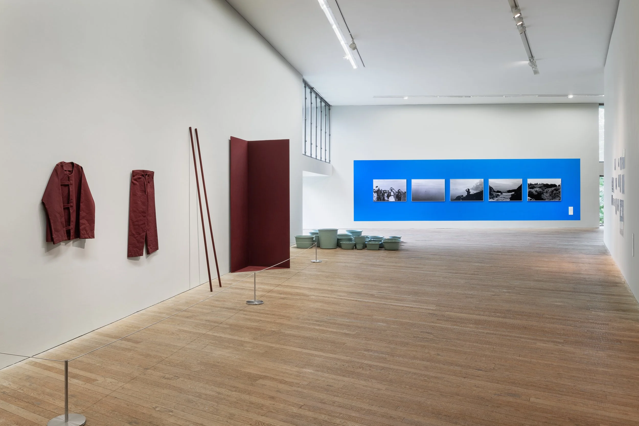 Art gallery interior with fashion items on the wall, ceramic pots on the floor, and a display of landscape photographs on a blue wall in the background.