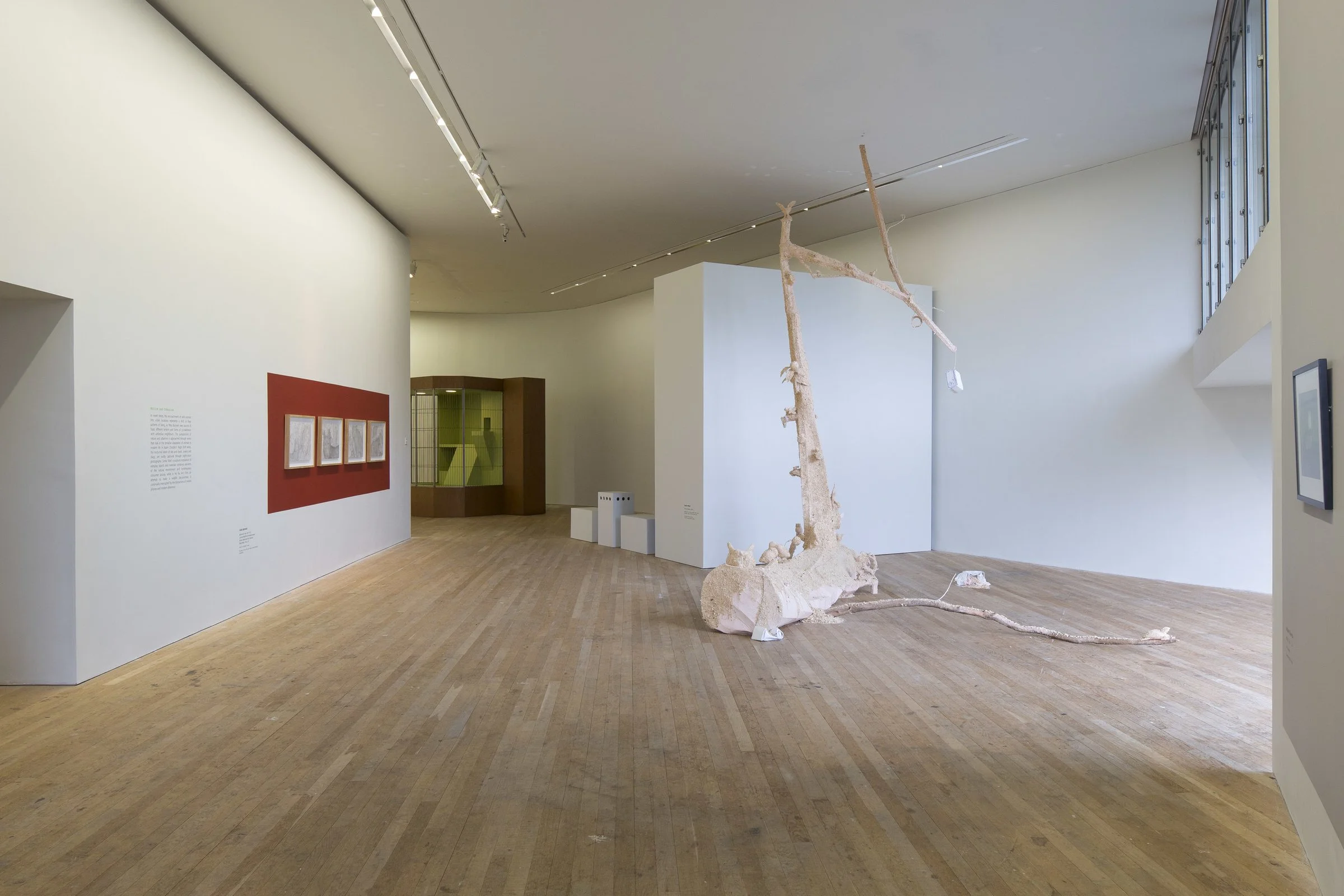 Art gallery with wooden floors, white walls, and natural light. Features a tall, thin, beige sculpture resembling a tree or branch, and framed artworks on the walls.
