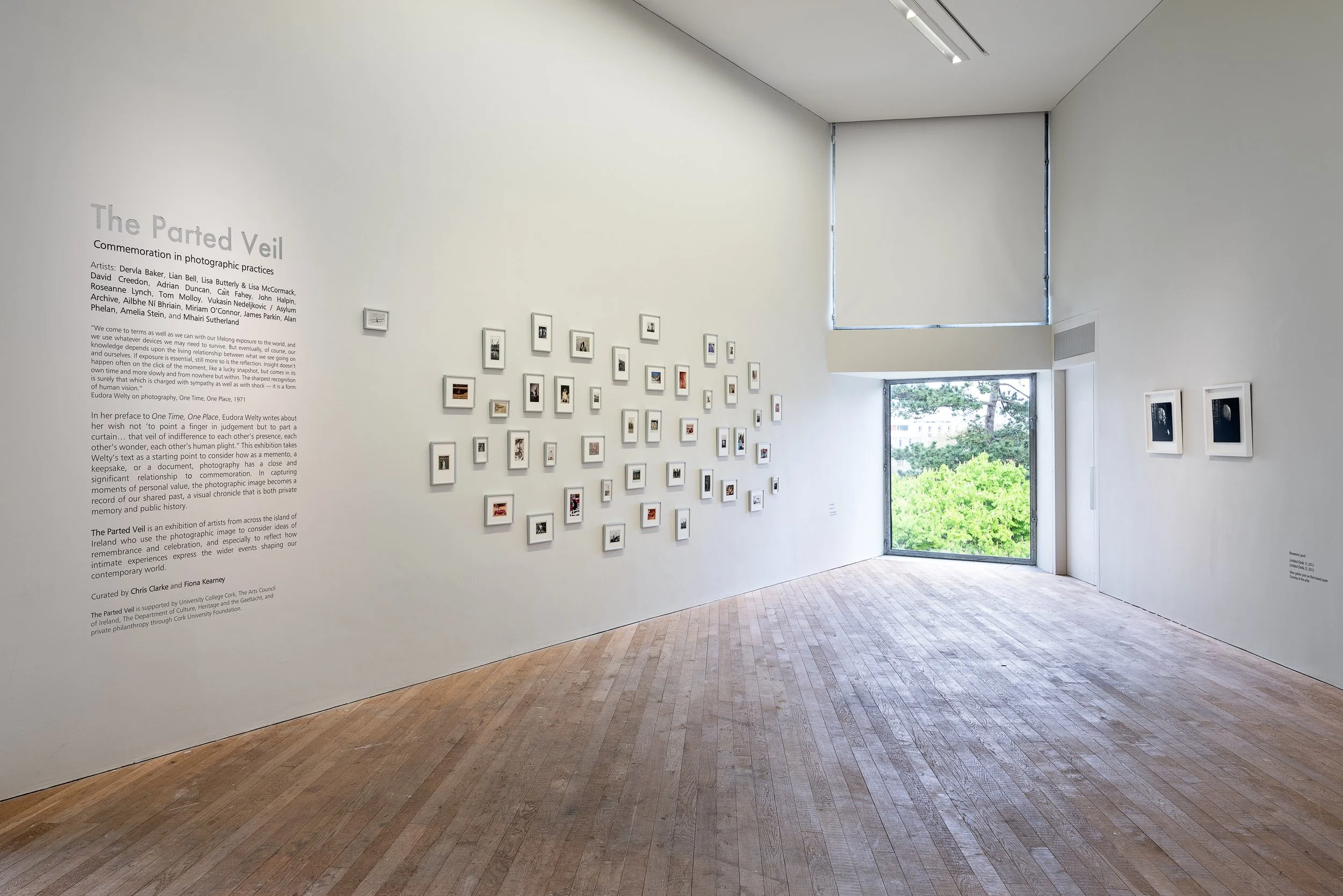 Interior view of an art gallery with white walls, wooden floor, and multiple small framed photographs displayed on the walls. A large window shows trees outside, and there is a text panel on the left wall about the exhibition titled 'The Parted Veil'