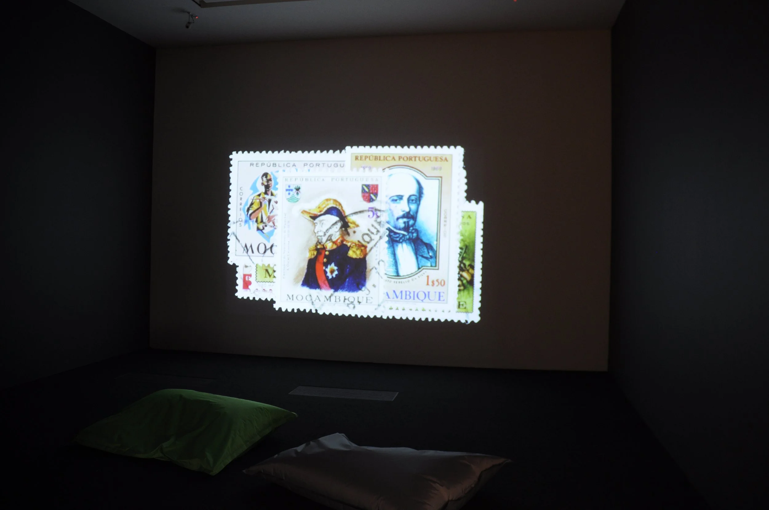 A projection of colorful postage stamps from Mozambique and Portugal is displayed on a dark wall in a room with two pillows on the floor, one green and one beige.