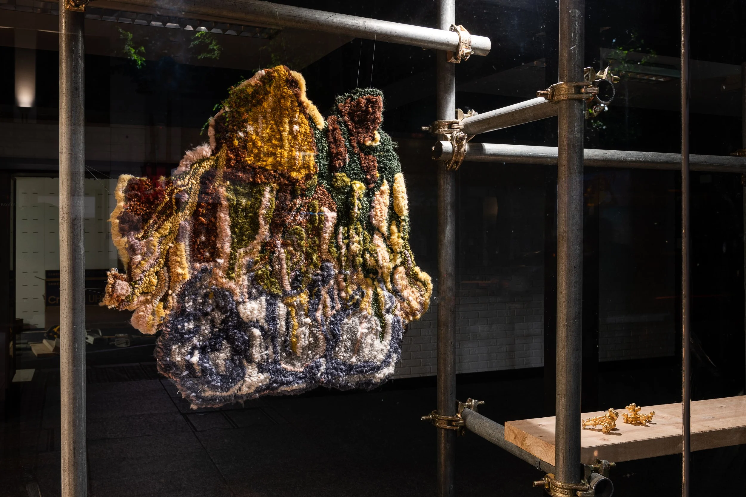Colorful textured handbag with abstract design displayed on a scaffolding structure, with a pair of gold earrings below it.