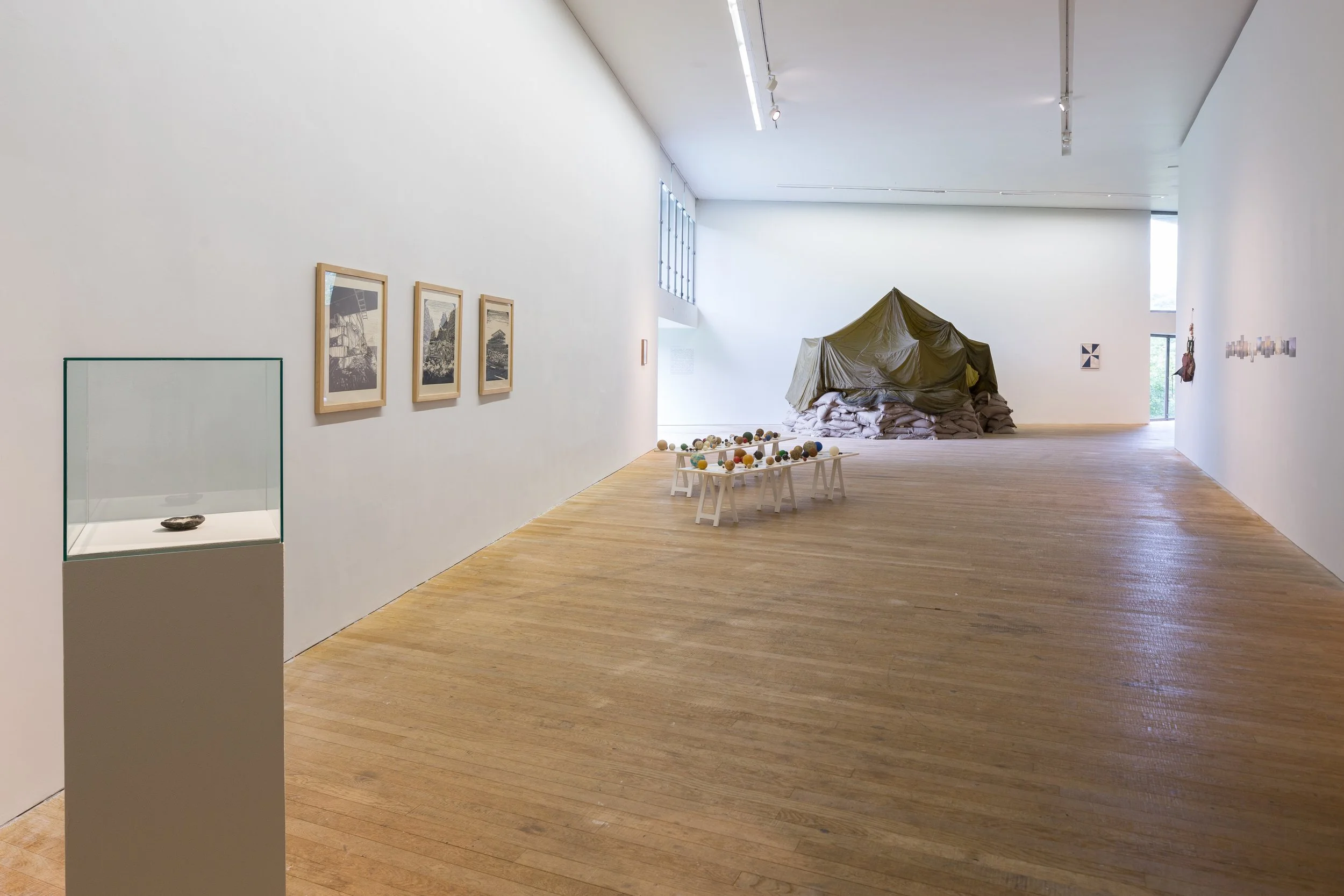 Modern art gallery with minimalistic white walls, wooden floor, framed black-and-white photographs, glass display case with a small artifact, and an abstract sculptural installation covered with a green tarp at the back.