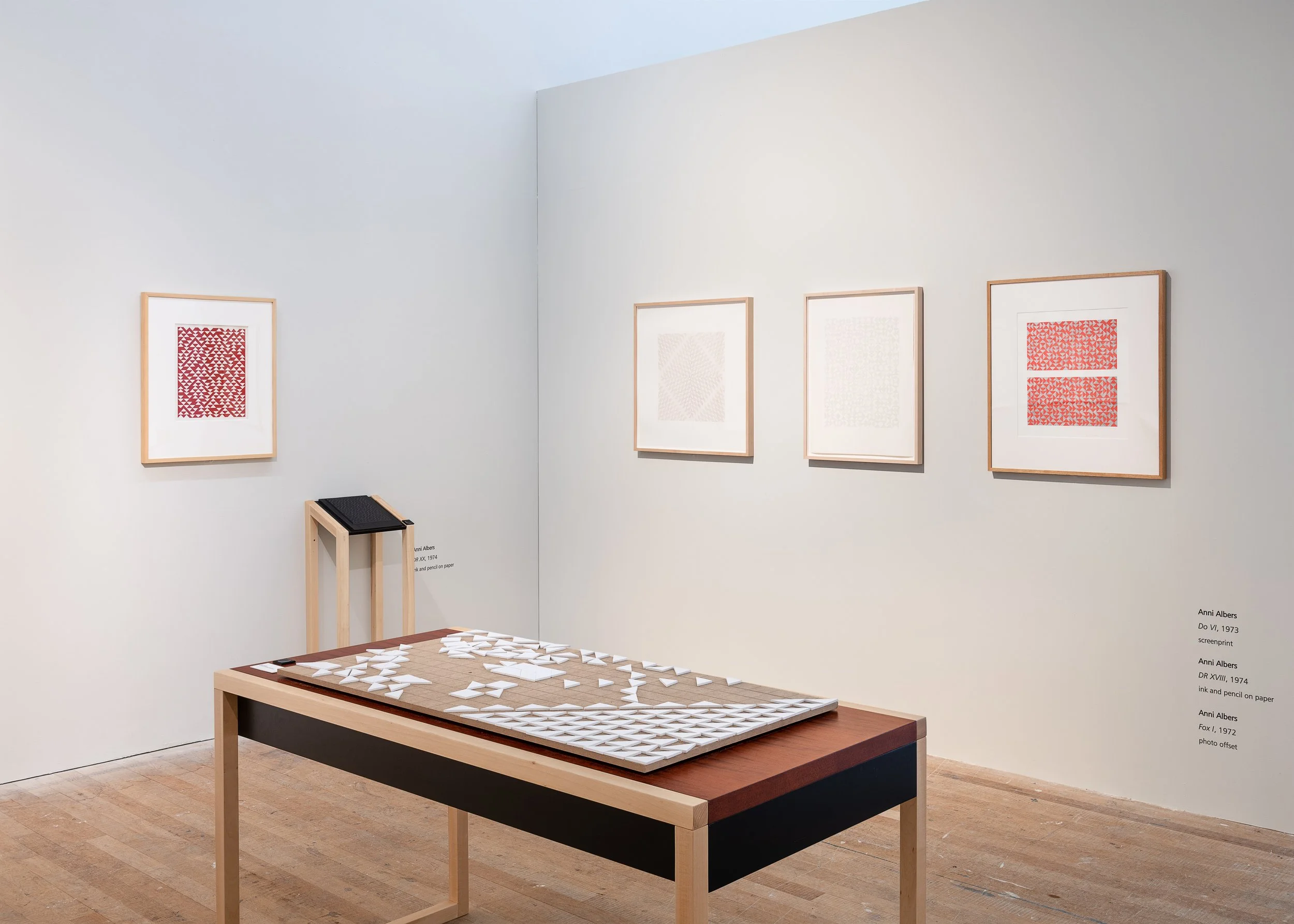 Art exhibit featuring framed geometric patterned prints on white walls, with a wooden table displaying white geometric paper sculptures, in a clean gallery setting.