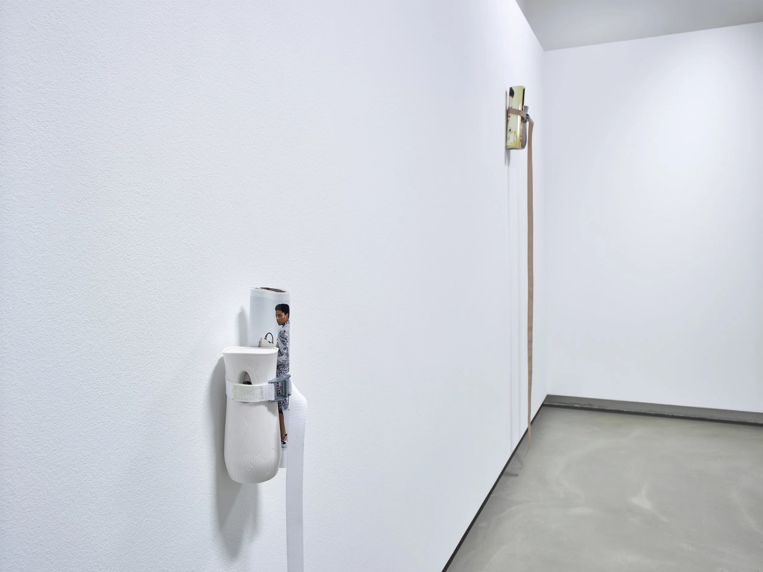 A white wall-mounted automatic hand sanitizer dispenser with a strap and a decorative picture of a woman. Behind it, a wooden wall-mounted device with a leather strap is visible in a minimalistic room with a plain gray concrete floor.