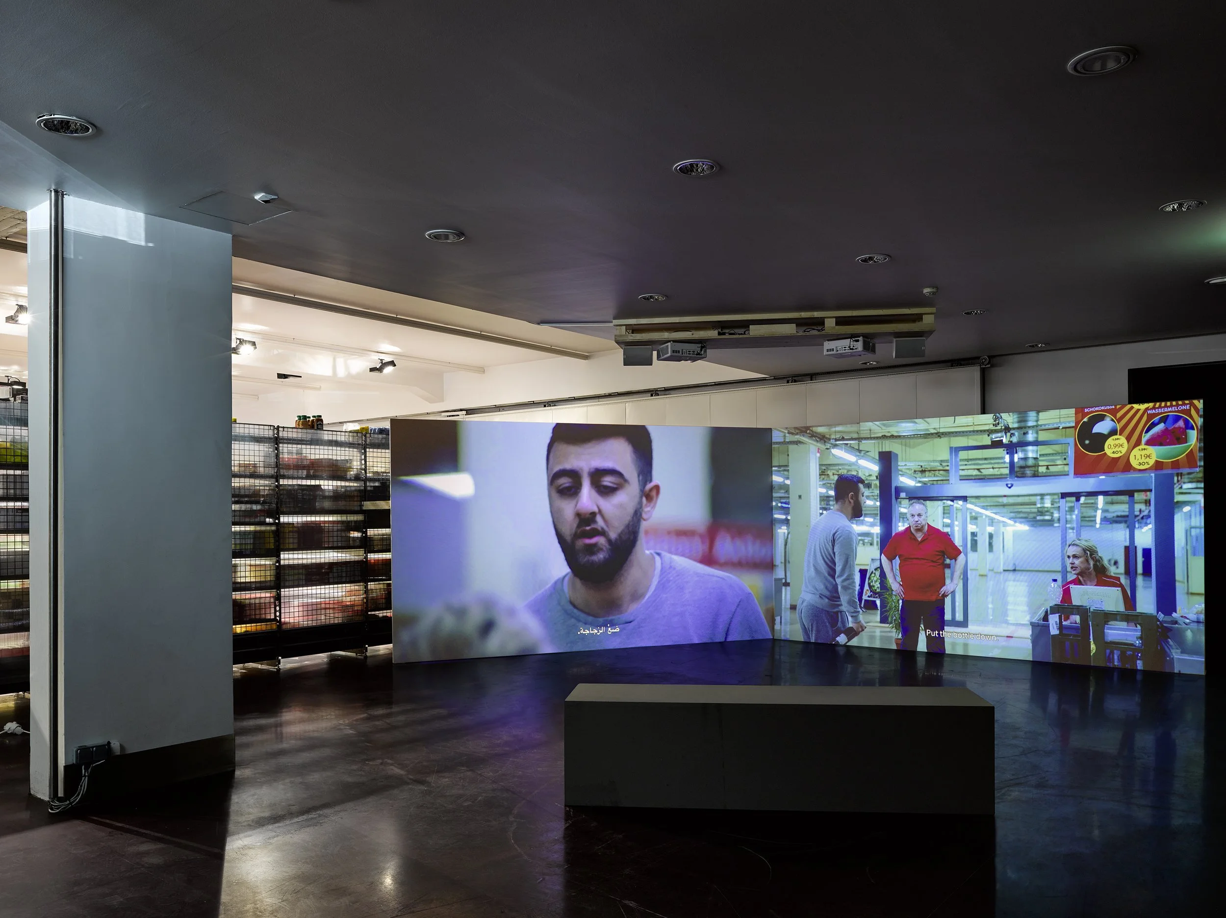 An indoor room with large electronic screens displaying a video of a man speaking. The room has dark flooring, white walls, and ceiling with multiple round recessed lights. There is a bench or low table in the foreground and shelves or storage units 