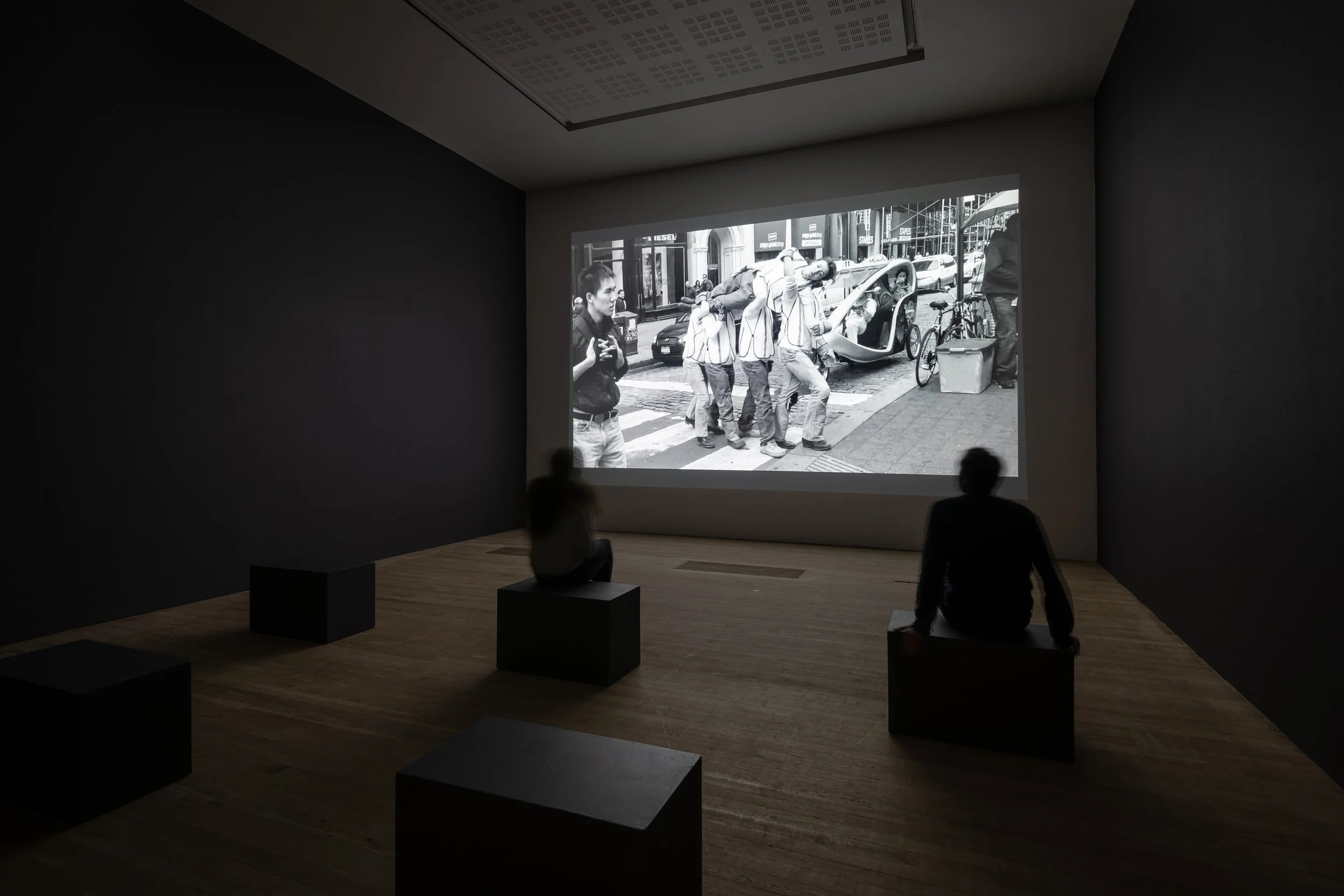 Two people watching a black and white photograph projected on a wall in a dimly lit room. The photograph shows a crosswalk with people walking, bicycles, cars, and city buildings.