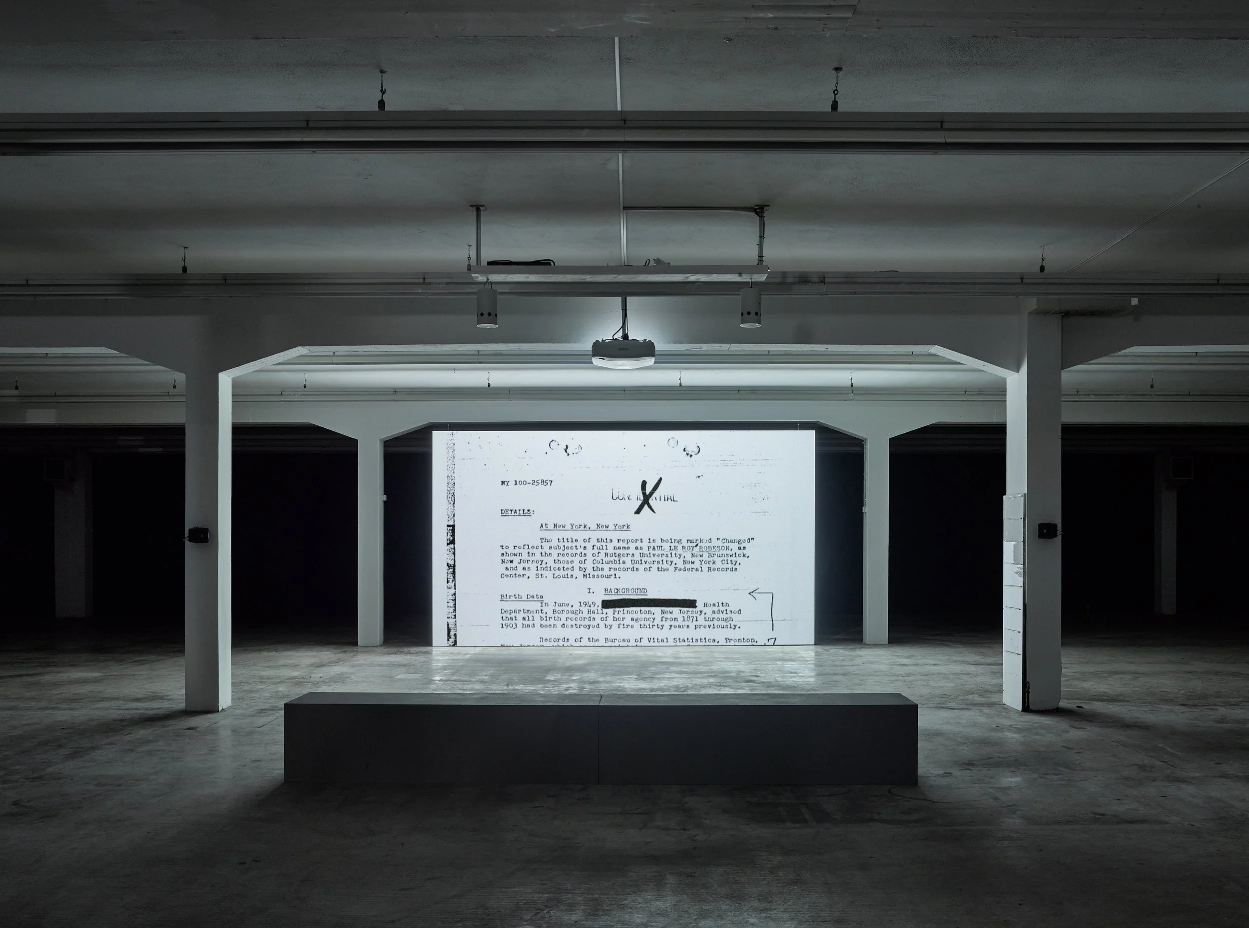 An art installation in a dimly lit warehouse or gallery featuring a large projected document on a wall with graffiti, and a short stage in front of it.