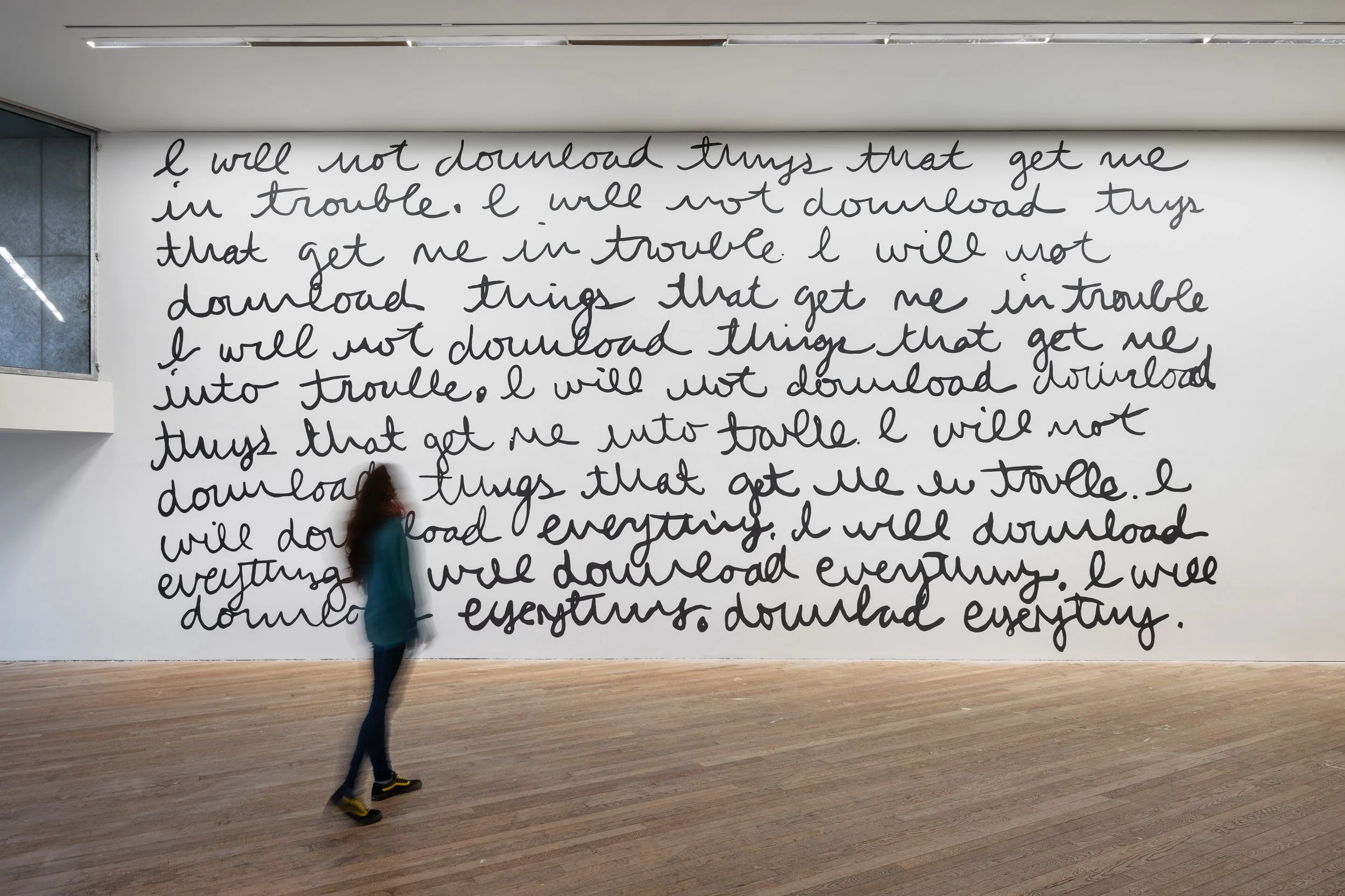 A woman walking past a large wall with handwritten text that repeats the phrase "I will not download things that get me into trouble."
