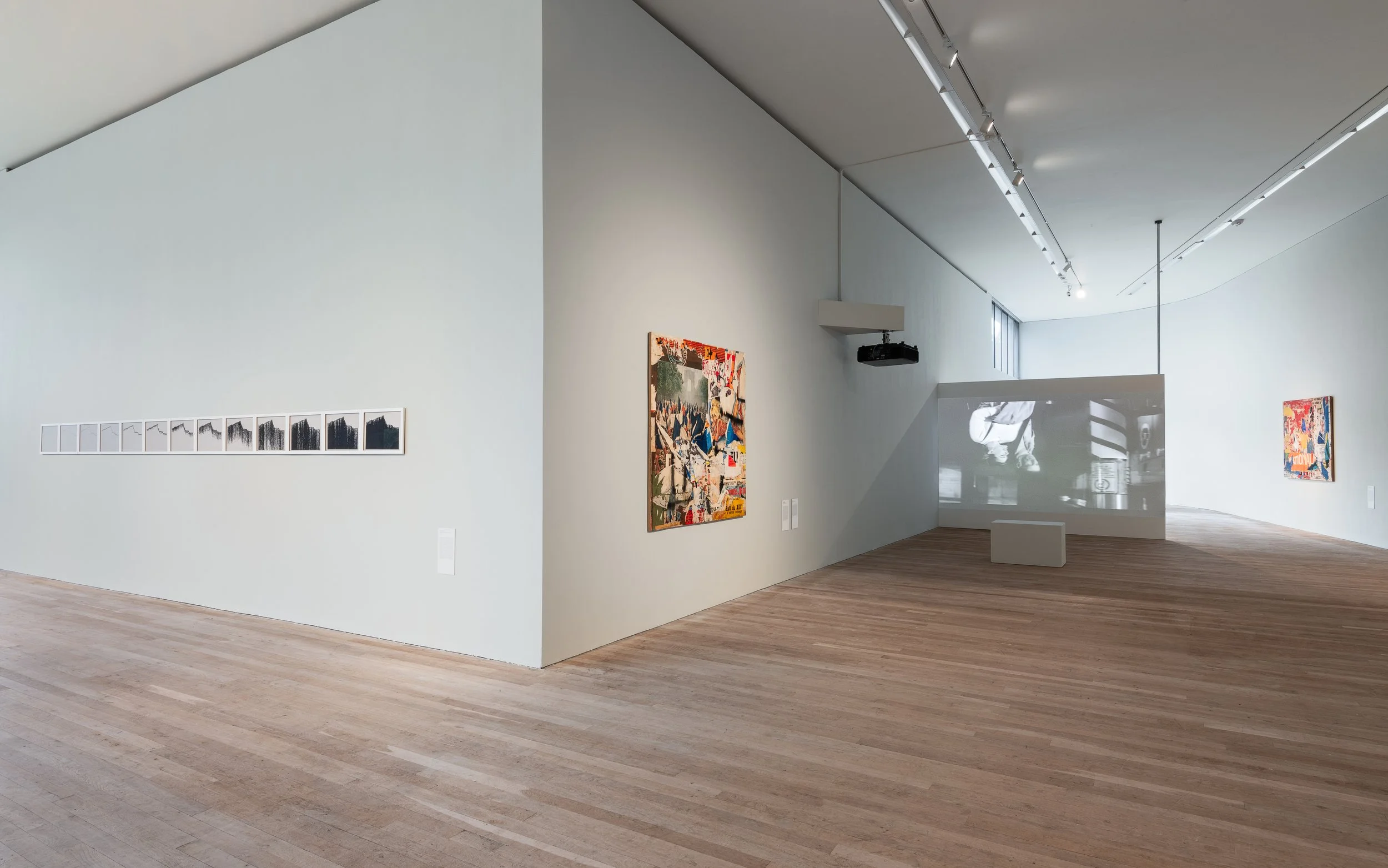 Modern art gallery with wooden floors, white walls, and multiple artworks displayed, including paintings and a video projection.