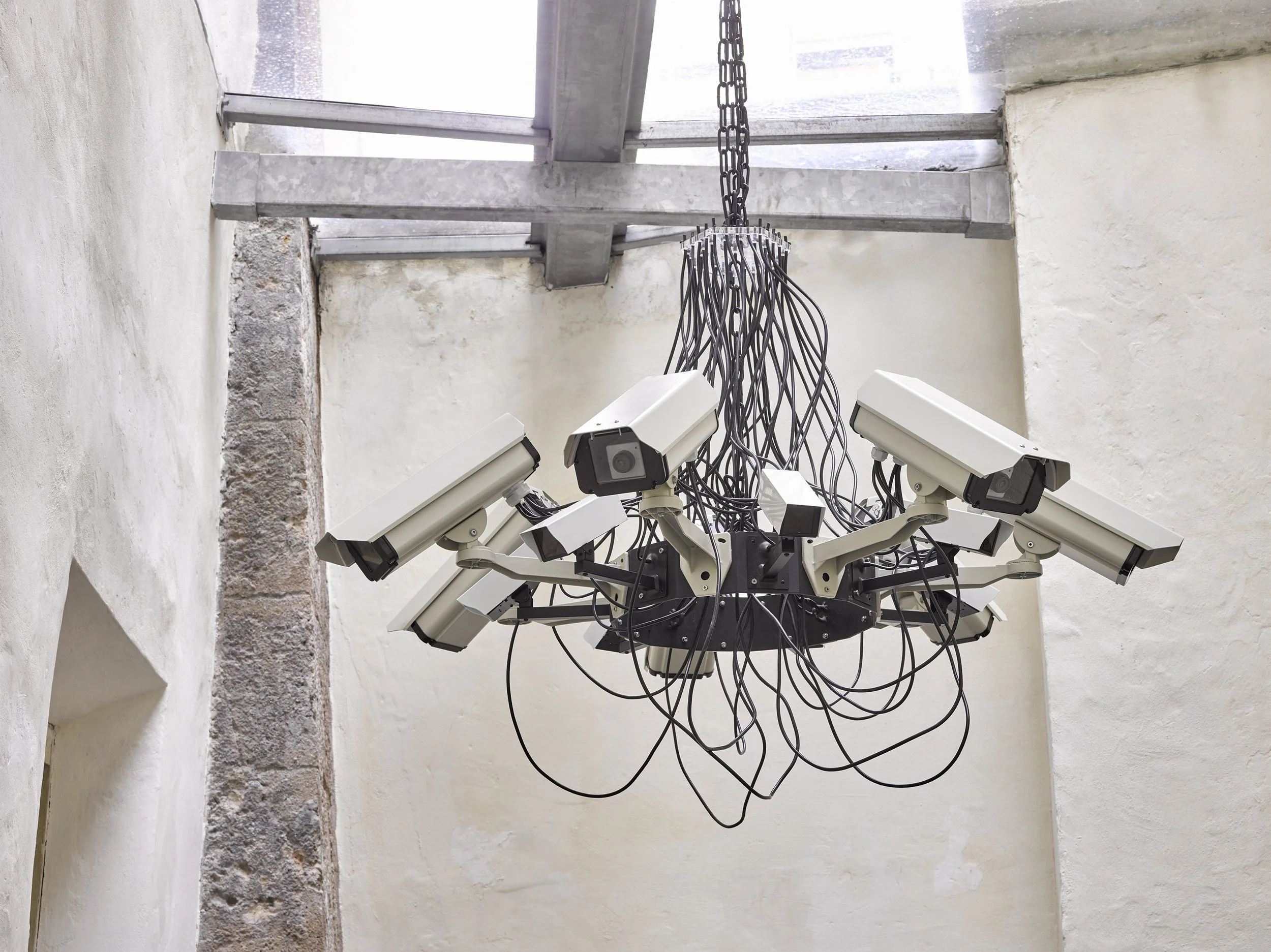 Interior view of a modern chandelier made of multiple surveillance cameras, hanging from the ceiling of a minimalistic room with white stucco walls and a skylight.