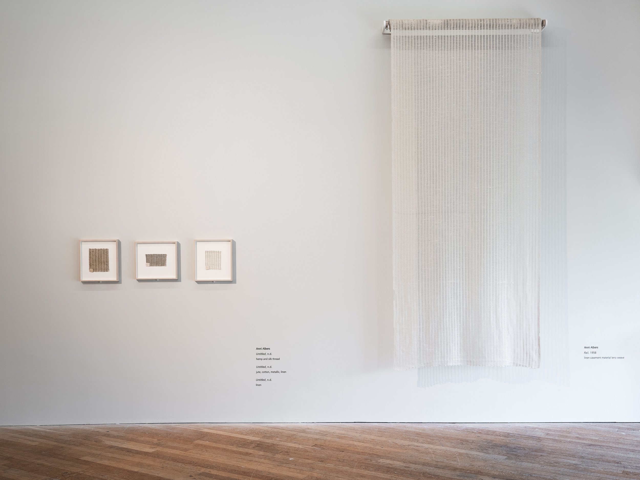 Art gallery wall displaying three framed textile pieces and a large hanging textile installation on a plain white wall.
