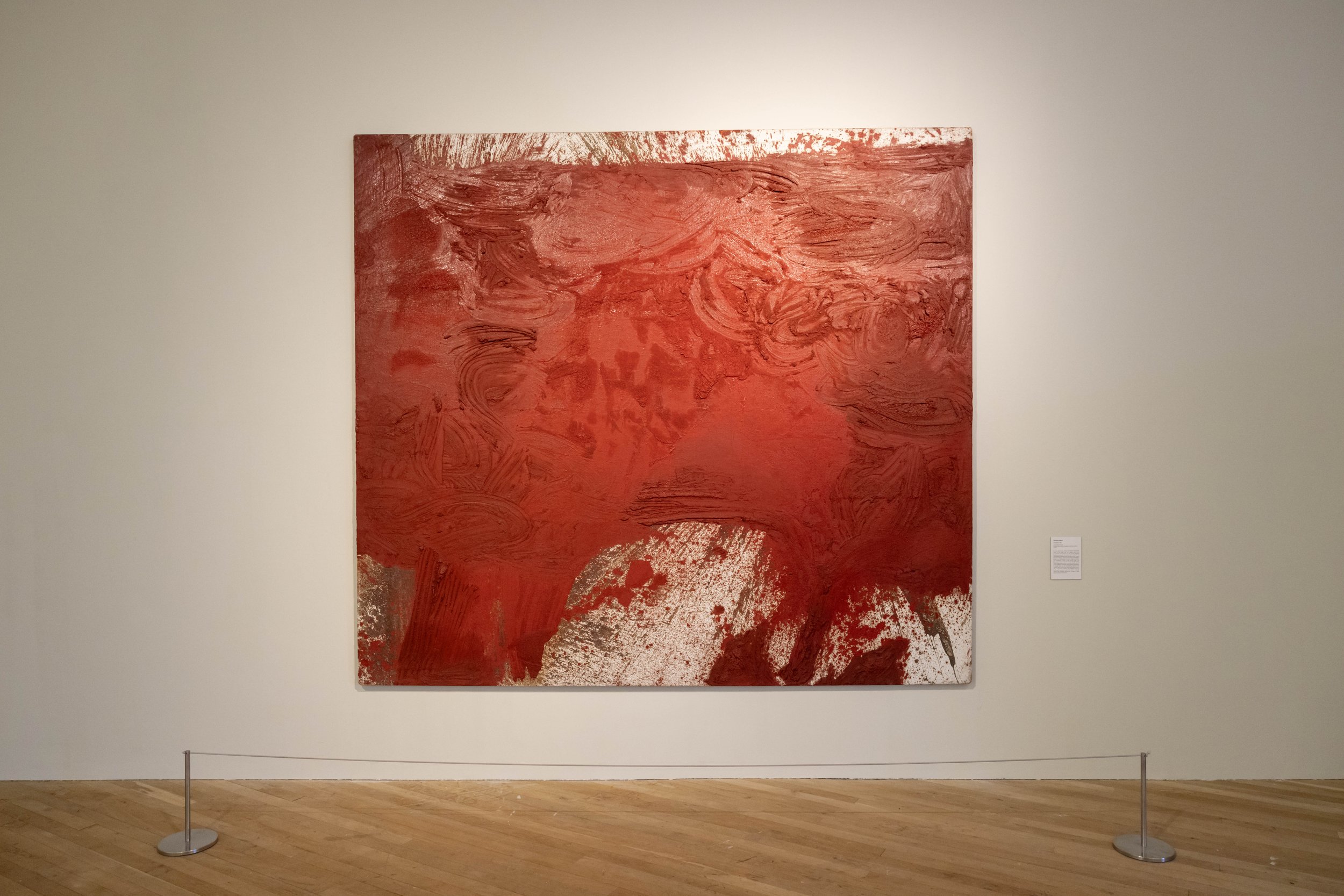 An abstract red painting with swirling patterns and textures, displayed on a gallery wall with a light-colored background.