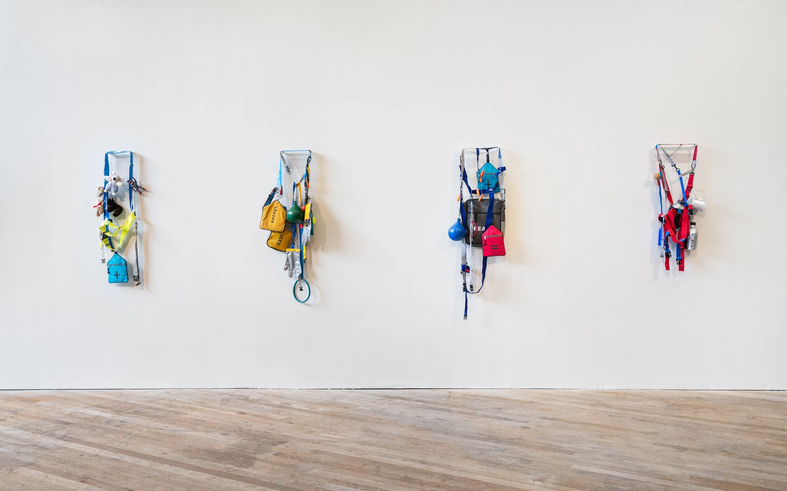 Four harnesses with climbing gear hanging on a plain white wall, labeled with color and containing various equipment like bags, ropes, and safety gear.