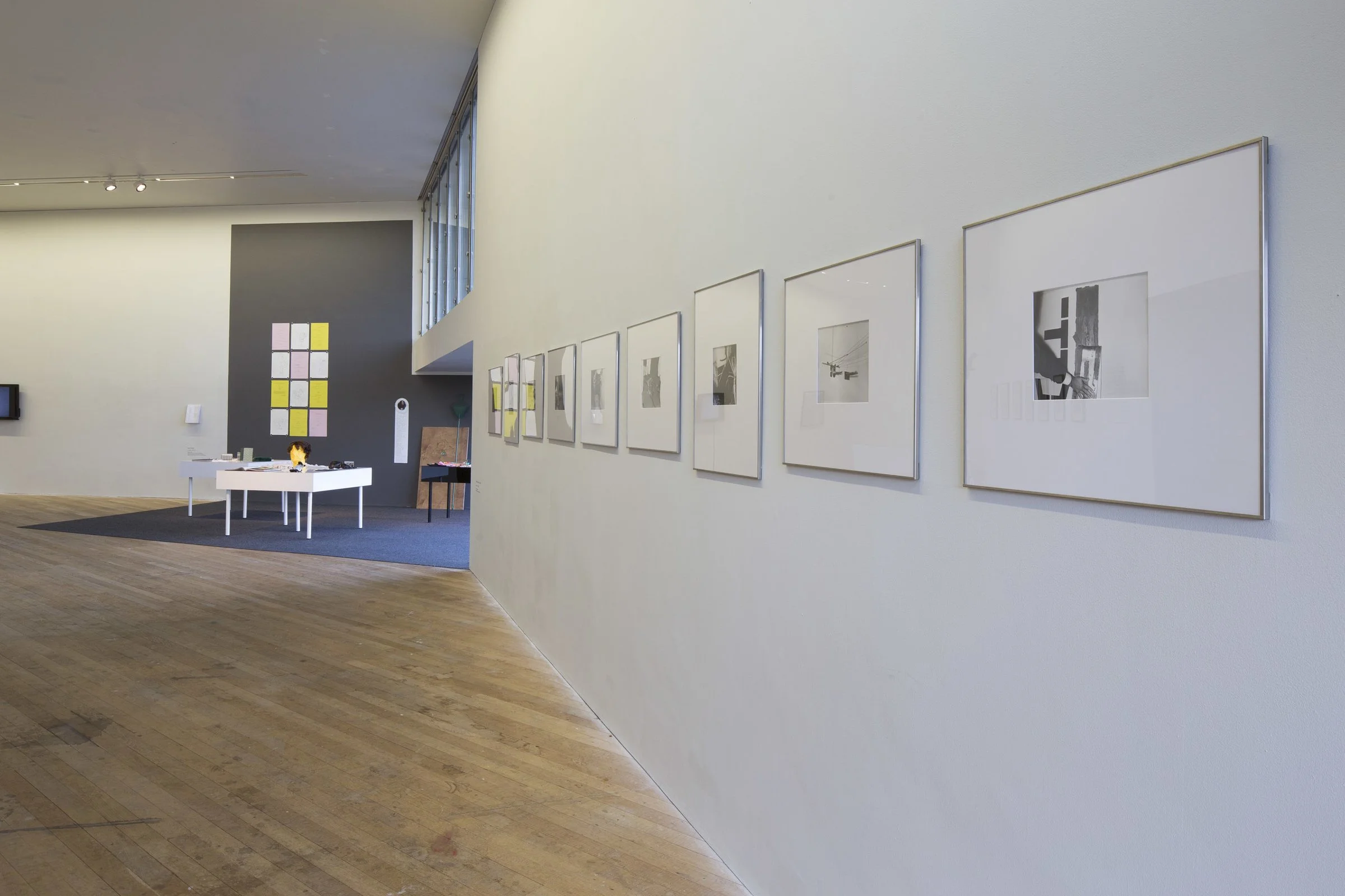 An art gallery with framed black-and-white photographs on a white wall and a display table with objects, featuring a dark accent wall with small framed art pieces and a large grid of colorful squares.