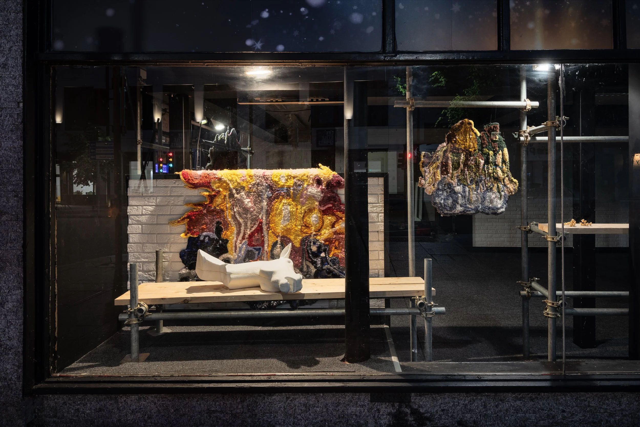 Art display in a storefront window with colorful mixed media sculptures and a white animal figure on a wooden platform.