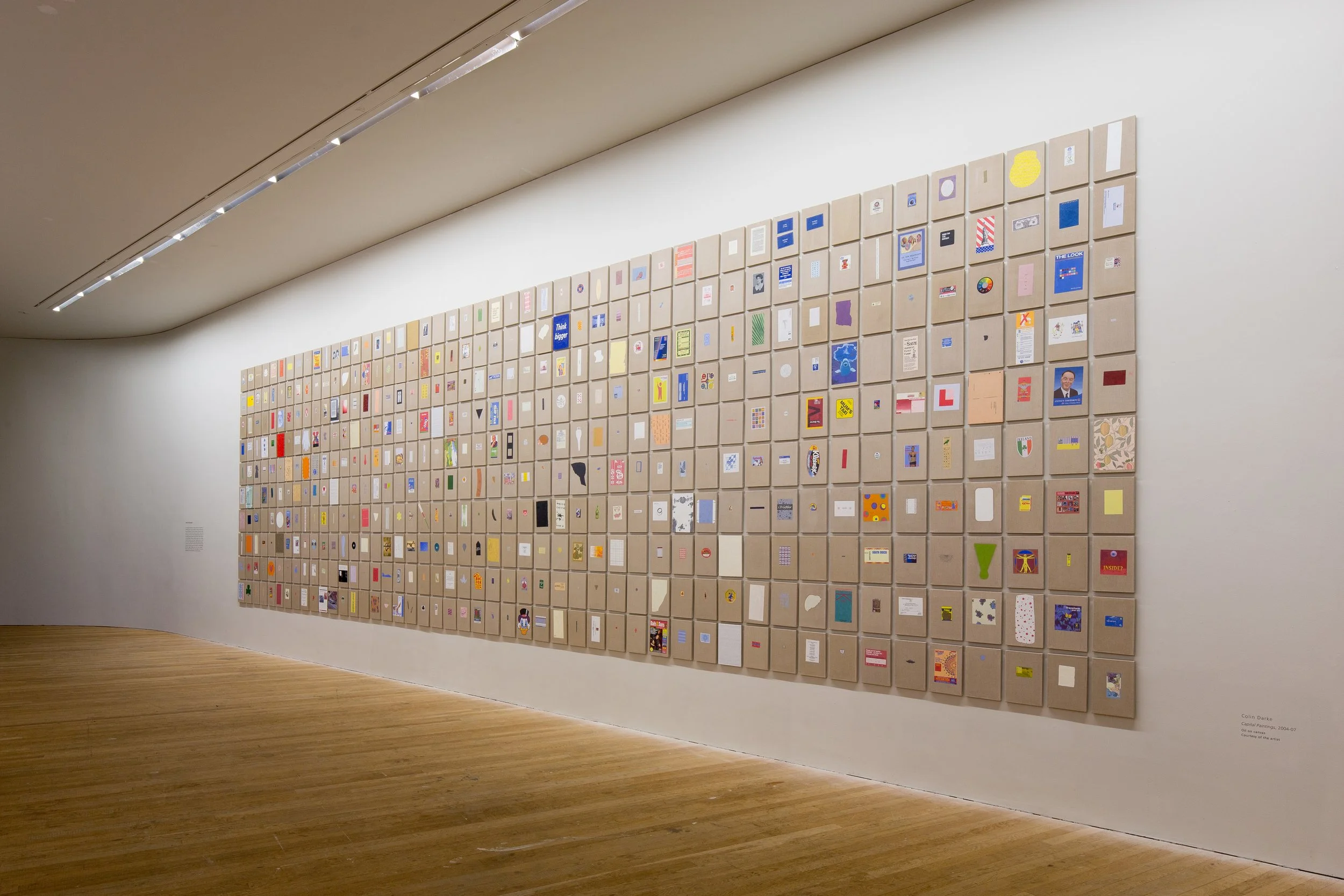 Large wall installation composed of many small framed pieces with colorful graphics, text, and patterns, arranged in a grid in an art gallery with wooden floors and white walls.